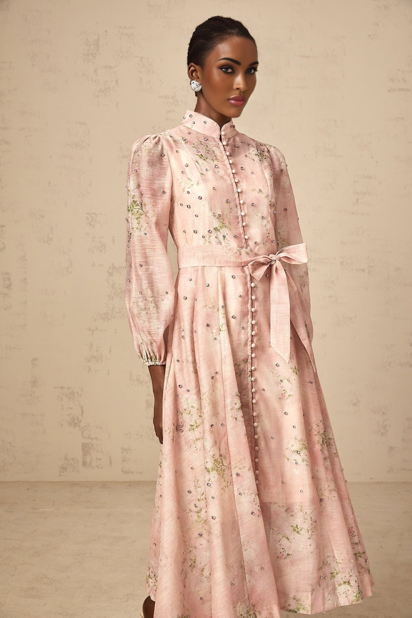 A woman wearing a pink midi dress with floral print and embellishments standing against a plain background