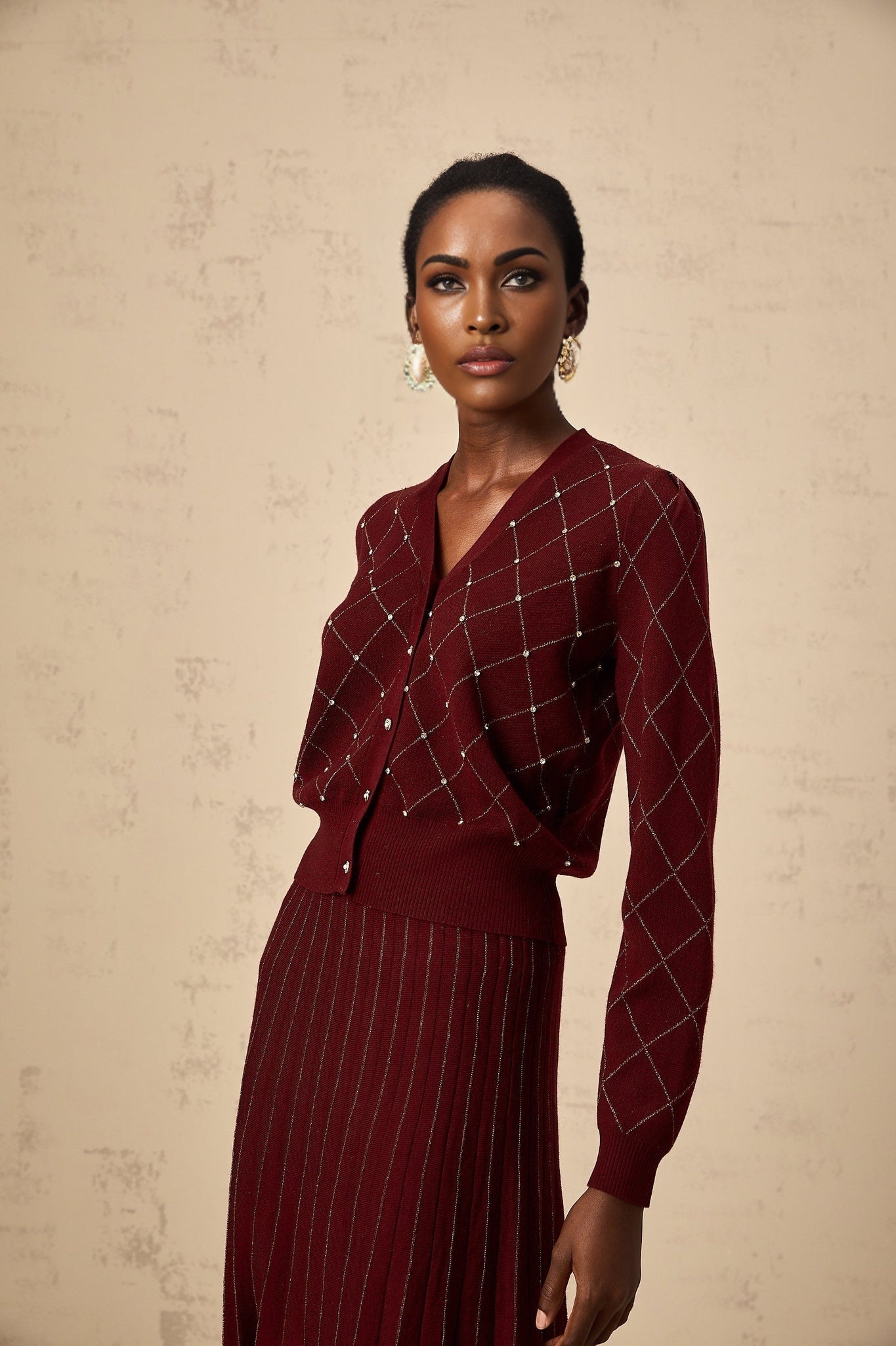 A woman wearing a red knit cardigan and matching skirt set featuring an argyle pattern with embellishments The cardigan has long sleeves and the skirt falls to knee length Both pieces are adorned with intricate intarsia knit designs