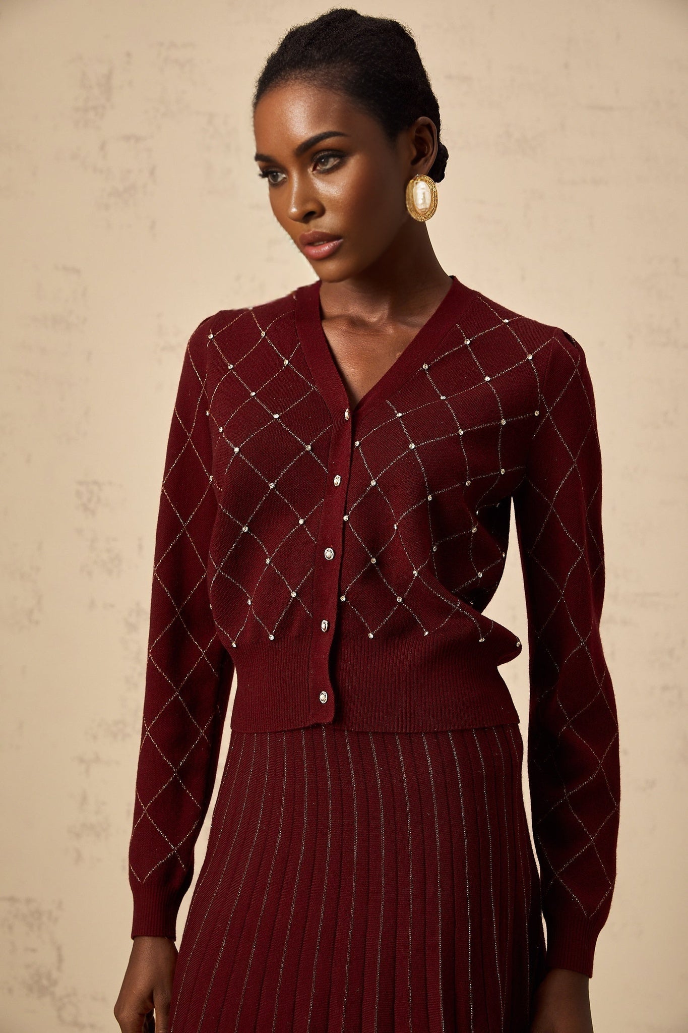 A woman wearing a red knit cardigan and matching skirt set with argyle pattern and embellishments The cardigan features long sleeves and buttons while the skirt falls to knee length Both pieces have an intricate intarsia knit design in shades of red