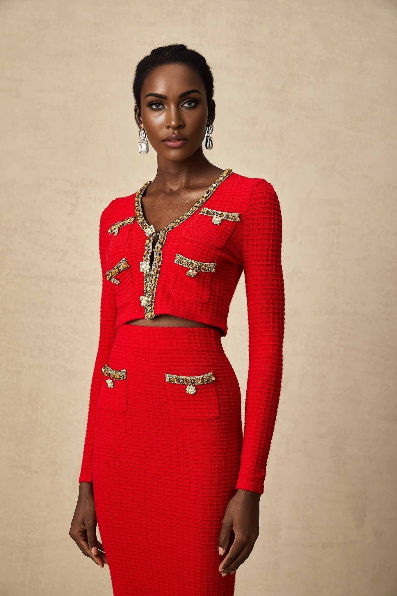 A woman wearing a red knitted cardigan and matching skirt set with embellished details The cardigan has long sleeves and the skirt falls to knee length Both pieces feature decorative embroidery or beading