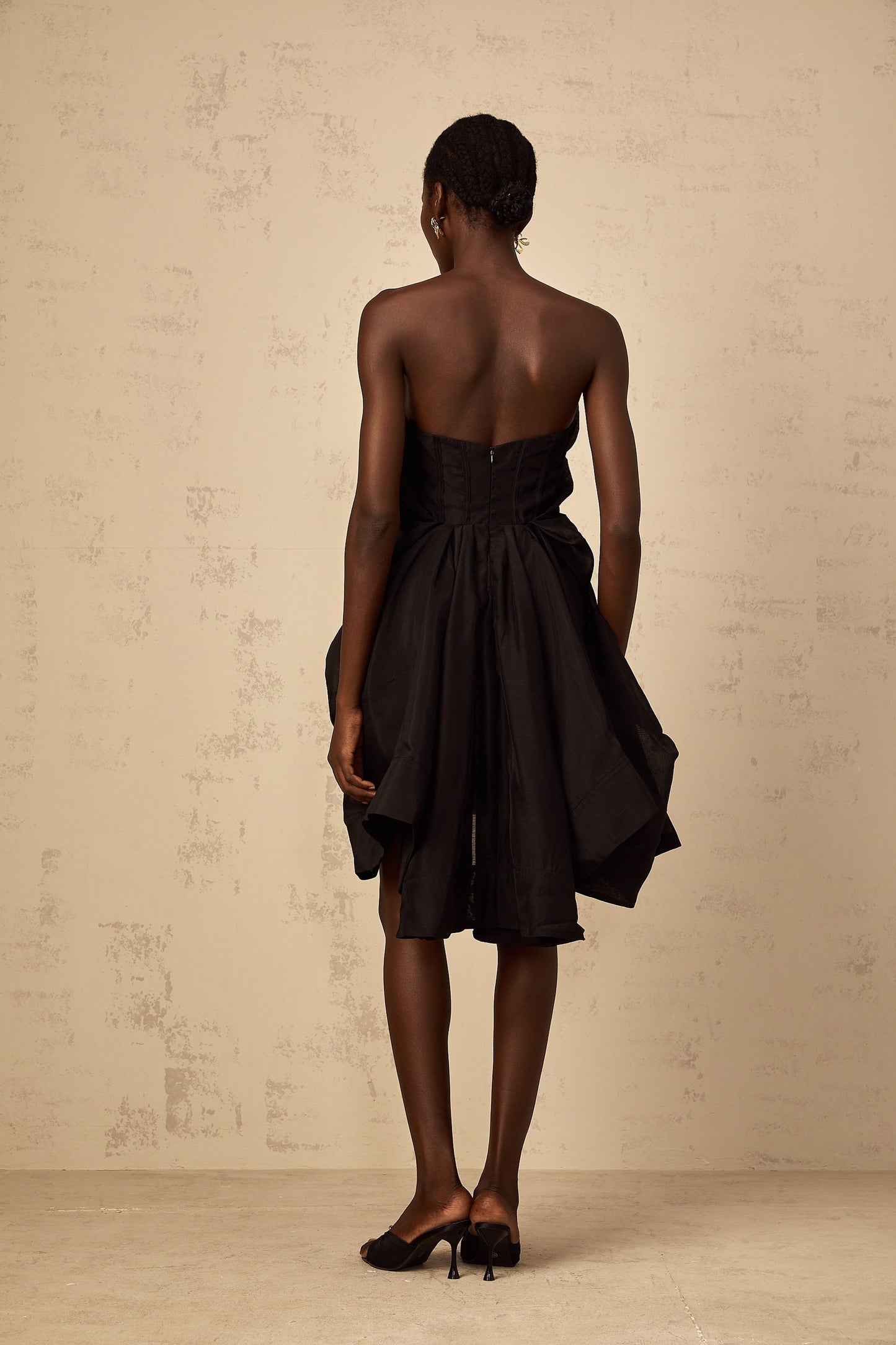 A woman wearing a short asymmetrical cocktail dress with pleated details standing in a relaxed pose against a plain background