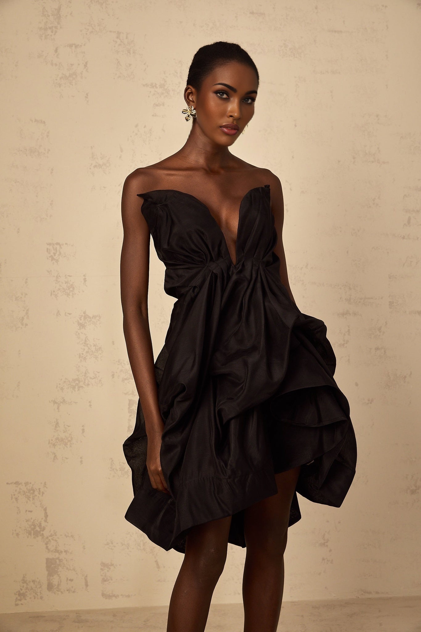 A woman wearing a short asymmetrical cocktail dress with pleated details The dress has one shoulder strap and falls above the knee The model is posed elegantly showing off the dresses unique design
