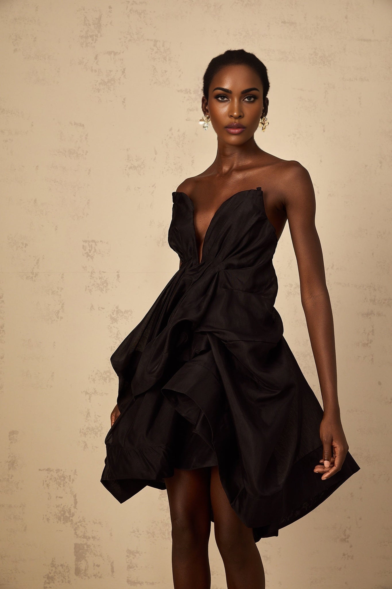 A woman wearing a short asymmetrical pleated cocktail dress called Elida The dress features a one shoulder design and falls above the knee with a fitted silhouette and flowing pleats