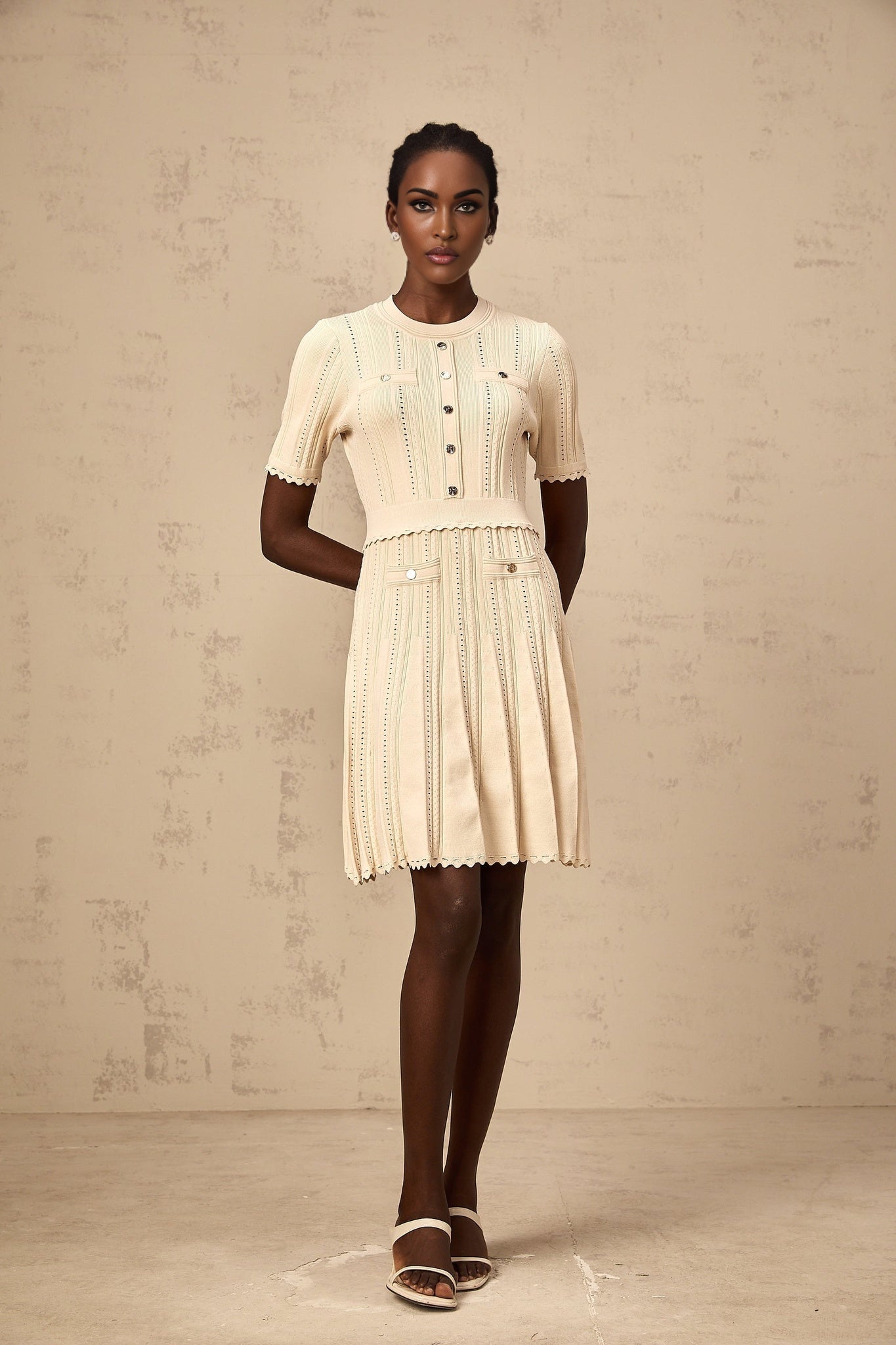 A woman wearing a short beige knitted dress with pleated skirt and delicate pointelle trim standing in a neutral background