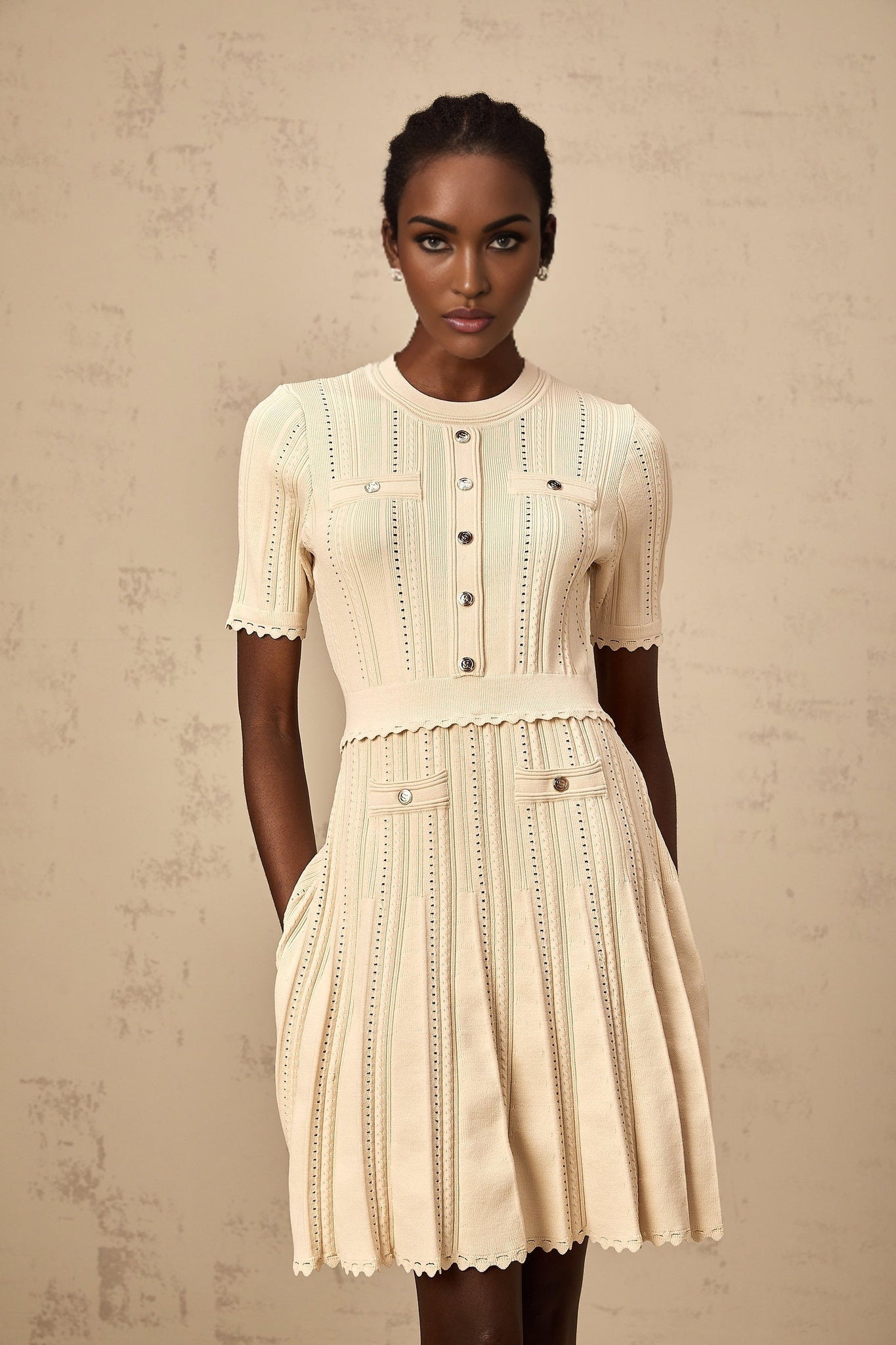A woman wearing a short beige knitted dress with pleats and delicate pointelle trim details standing in a neutral setting