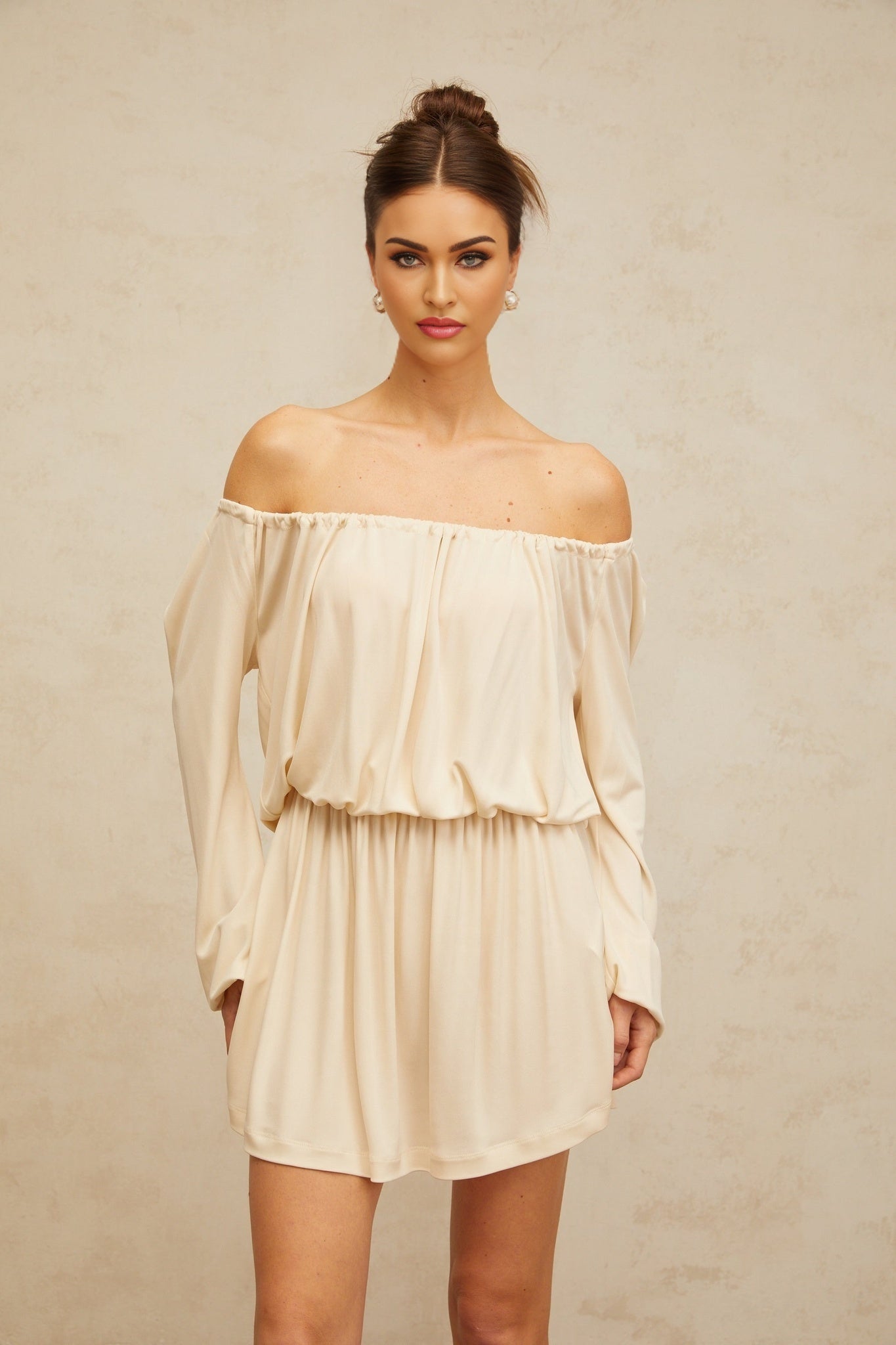 A woman wearing a short beige structured mini dress with a fitted silhouette and clean lines standing against a plain background