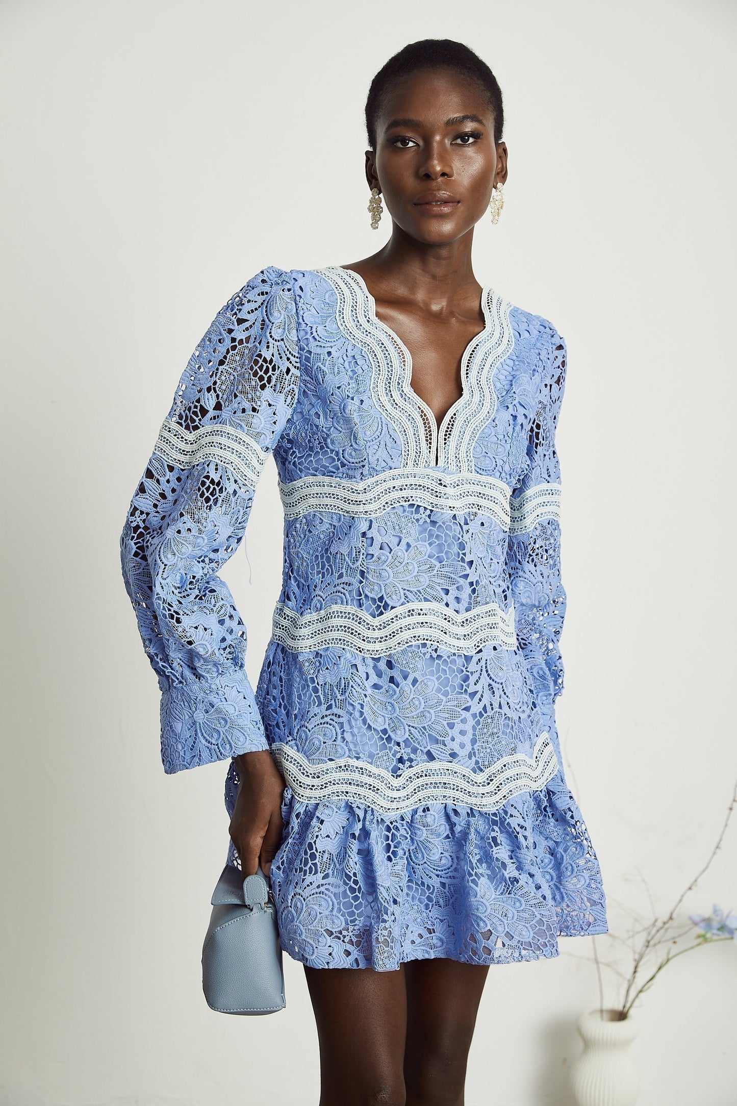 A woman wearing a short Gaelle dress with a V neckline and floral embroidery details standing against a plain background