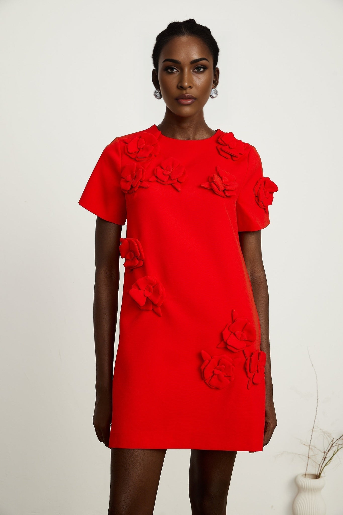 A woman wearing a short red mini dress with floral details and short sleeves standing against a plain background