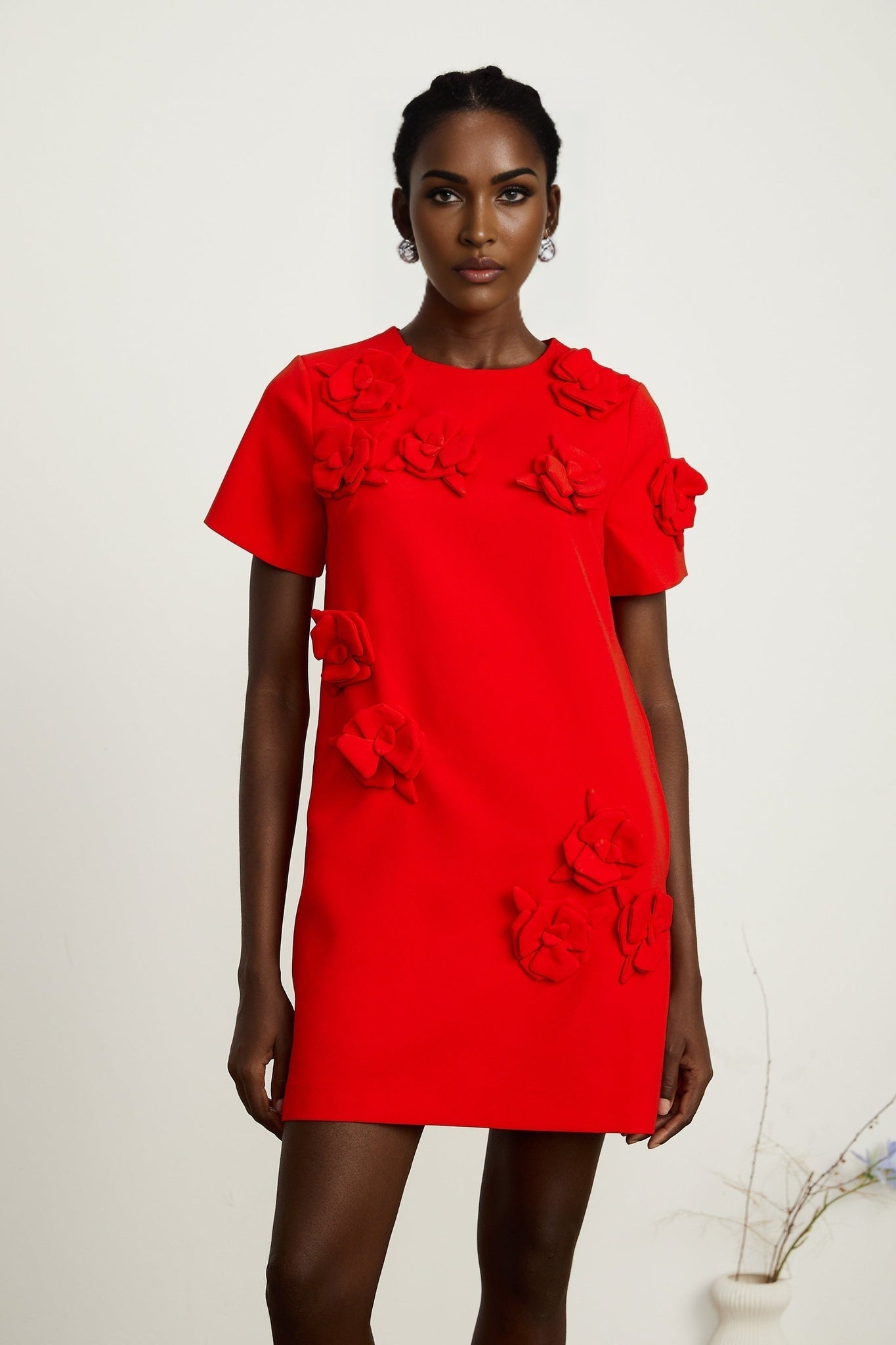 A woman wearing a short red mini dress with floral details and short sleeves standing against a plain background