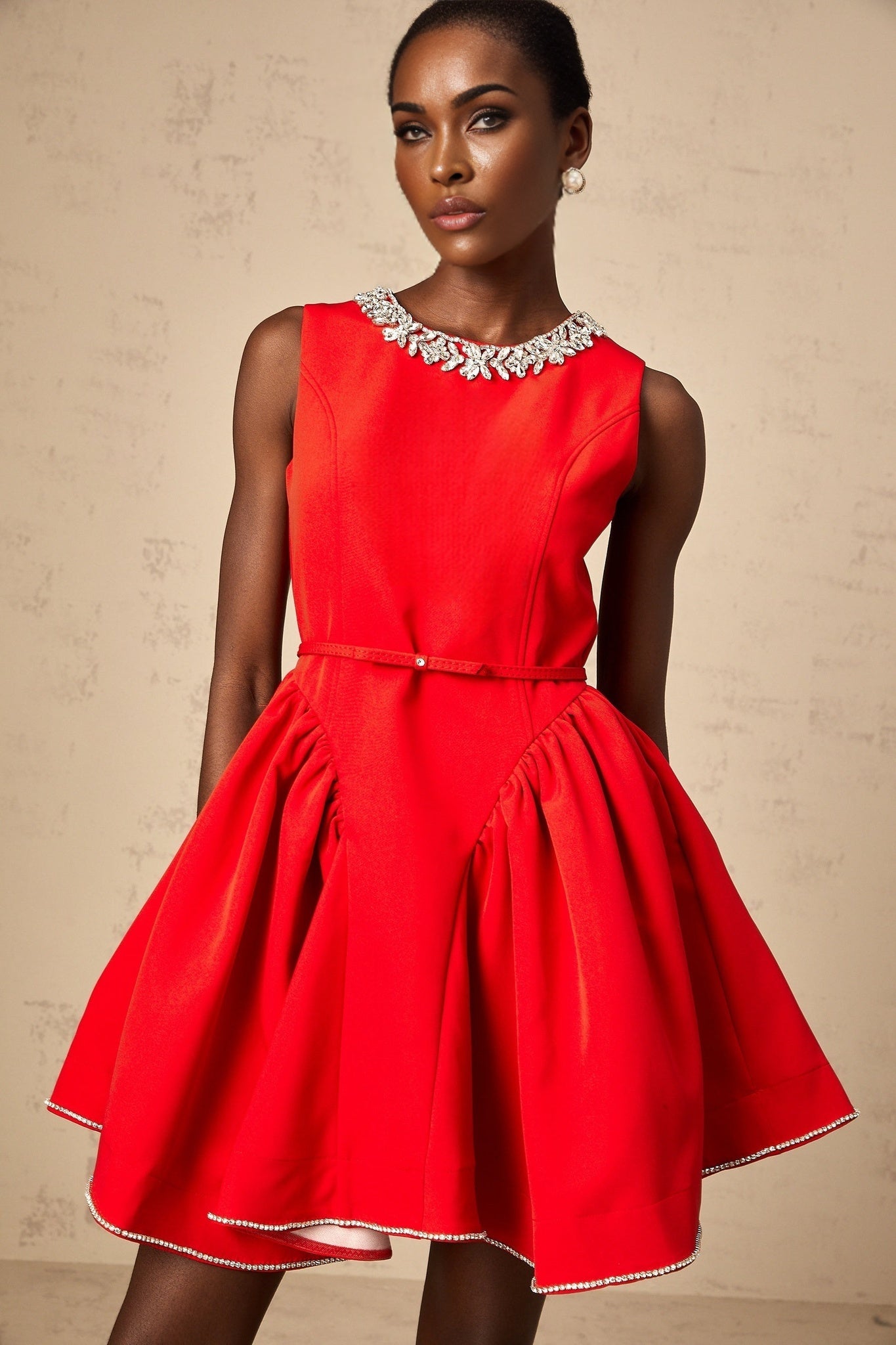 A woman wearing a short red pleated dress with embellishments standing against a plain background