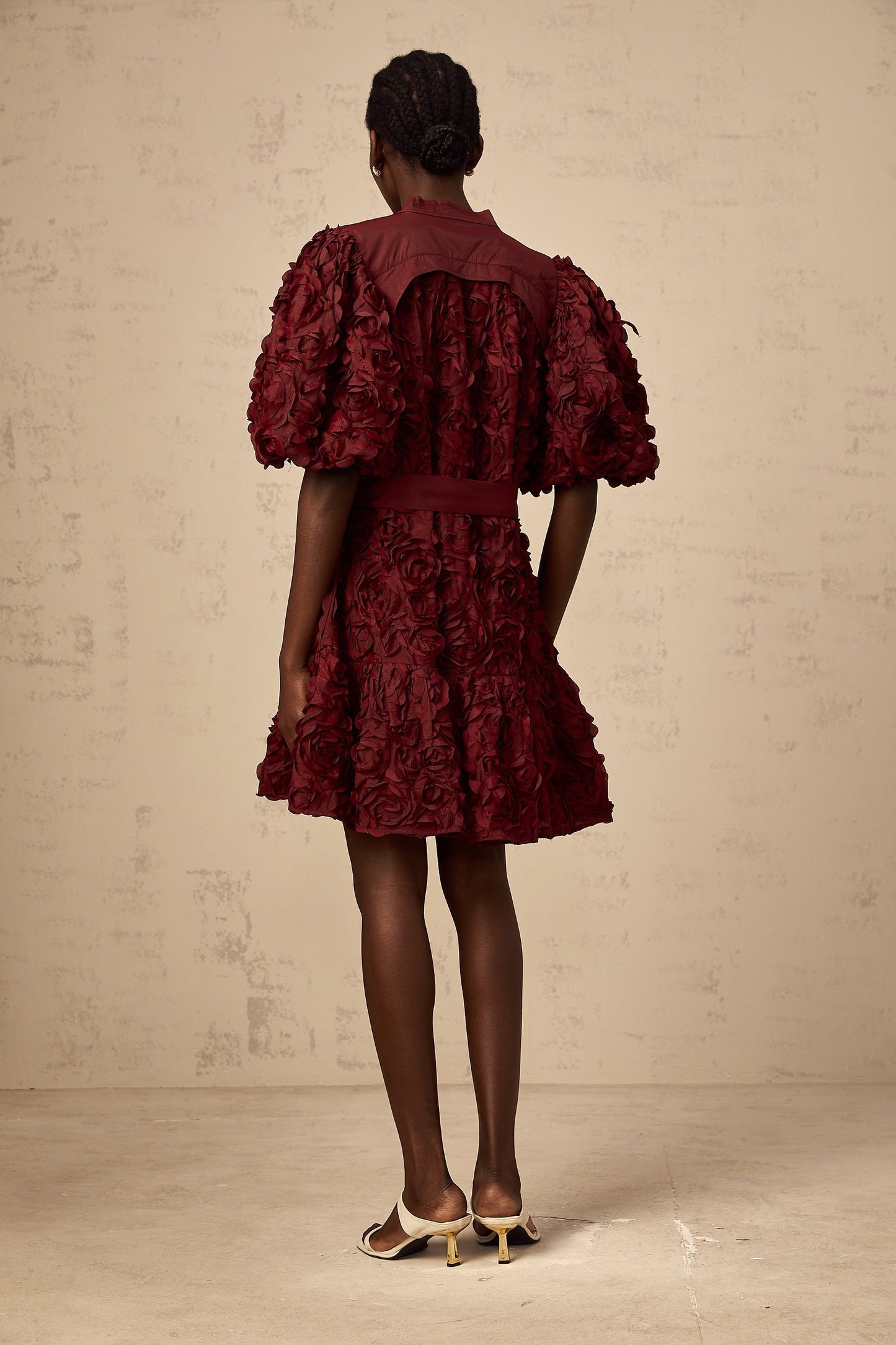 A woman wearing a short winered dress with textured 3D petal details and puffed sleeves standing against a plain background
