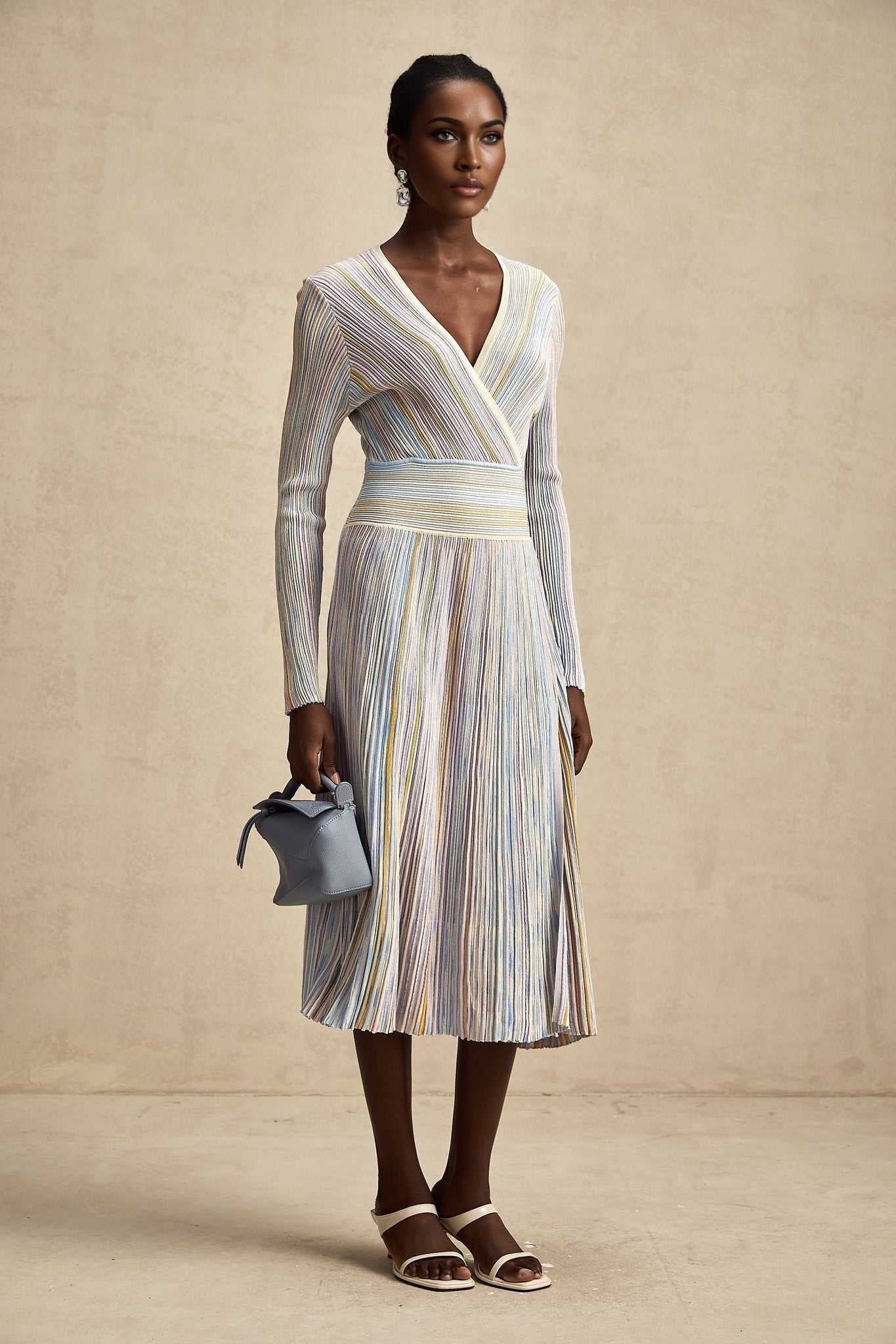 A woman wearing a vertical striped knitted midi dress in multiple colors with long sleeves and a high neckline standing against a plain background