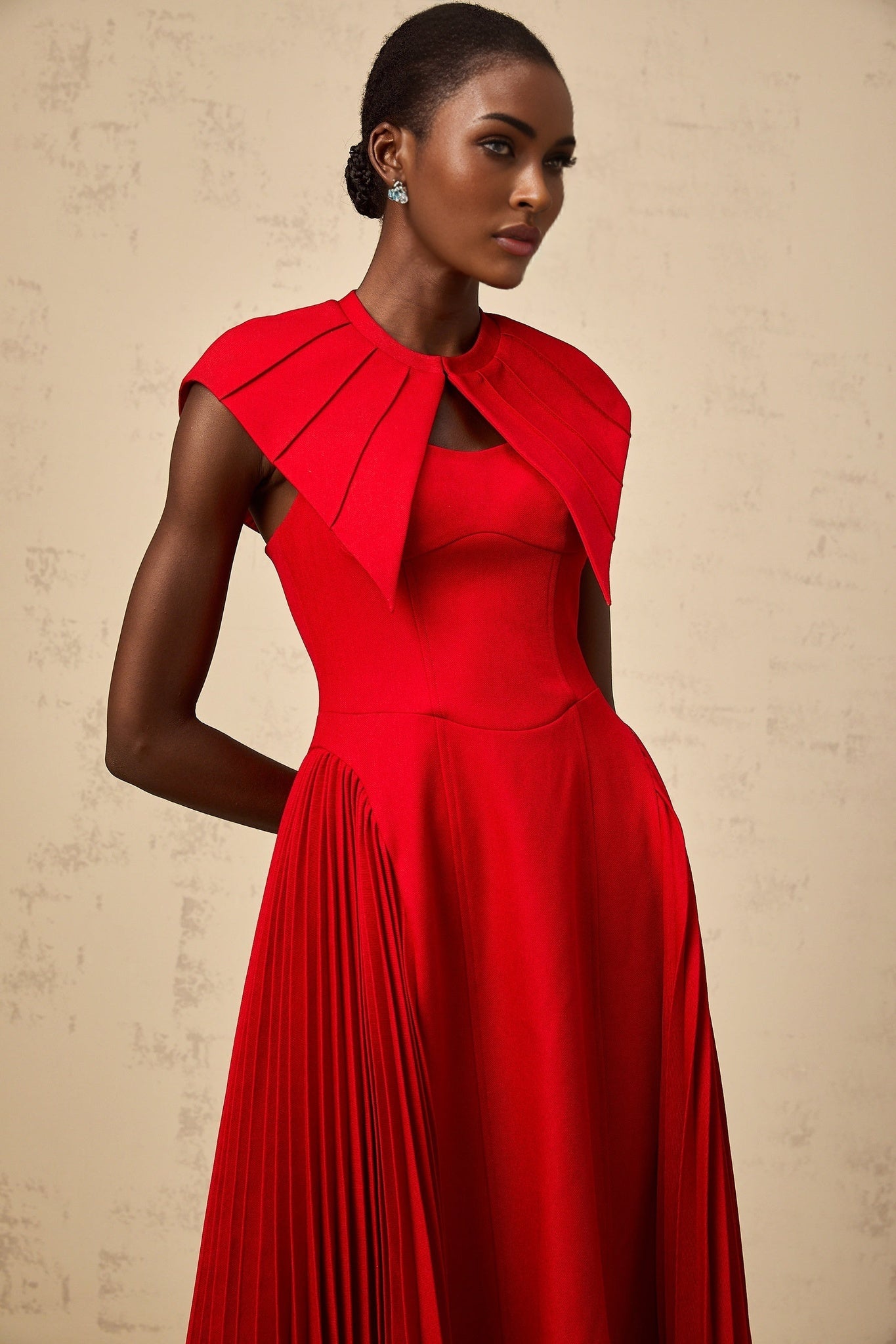 A woman wearing a vibrant red midi dress with a capelike design and pleated skirt standing against a plain background