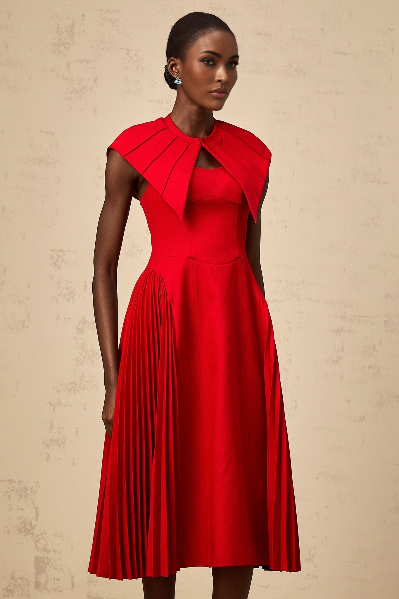 A woman wearing a vibrant red midi dress with a capelike design and pleated skirt standing in a neutral background