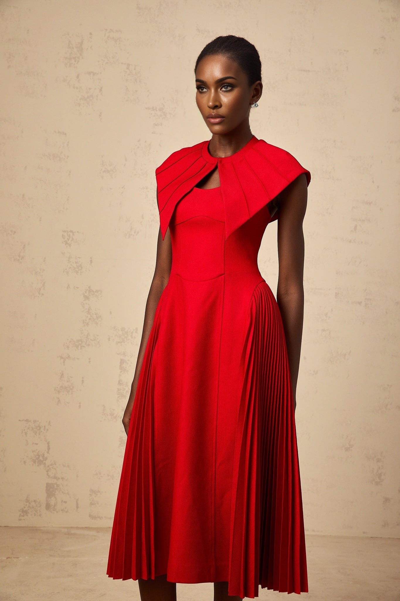 A woman wearing a vibrant red midi dress with a capelike design and pleated skirt standing in a neutral background