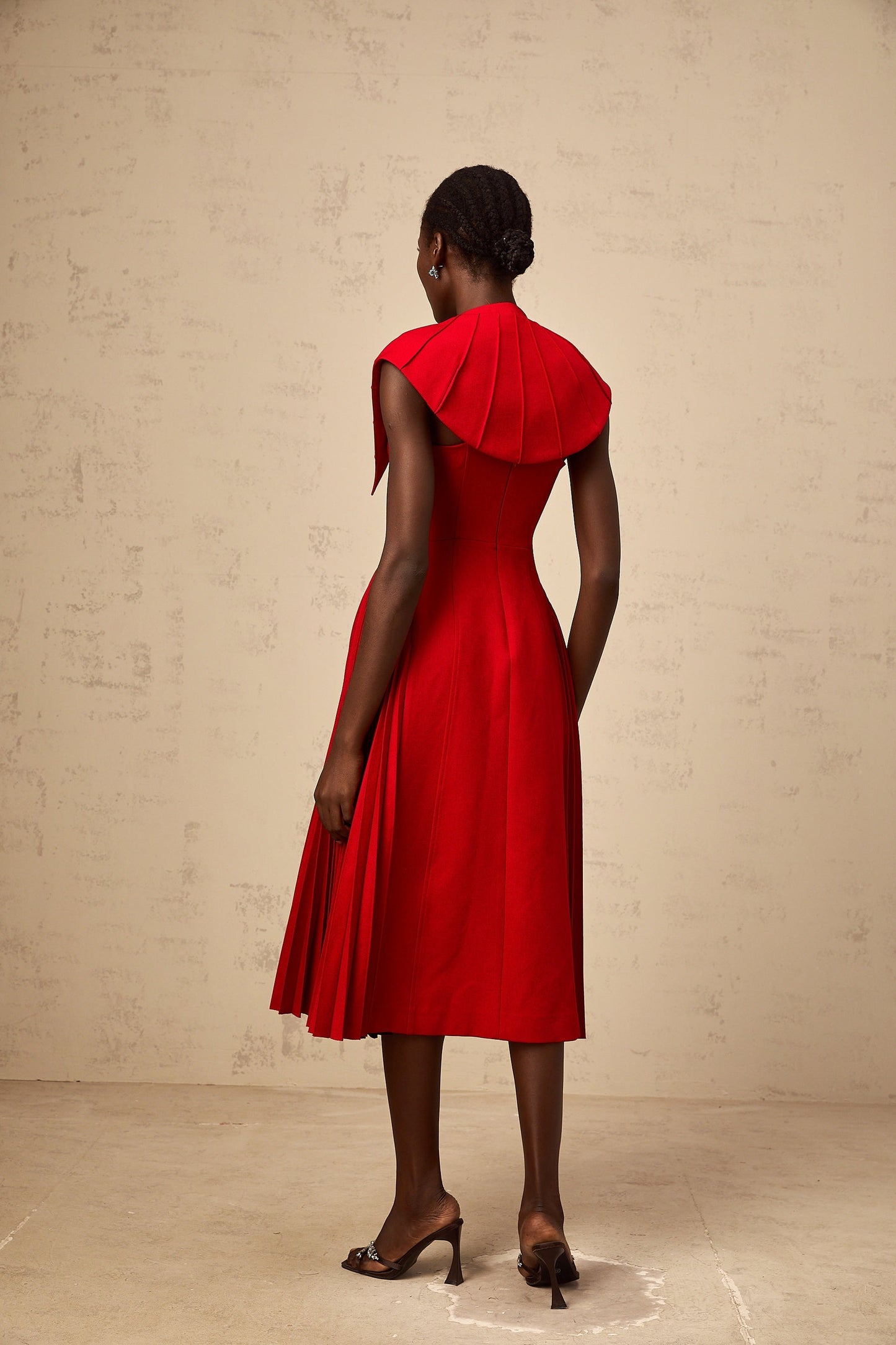 A woman wearing a vibrant red midi dress with a capelike design and pleated skirt standing in a neutral setting