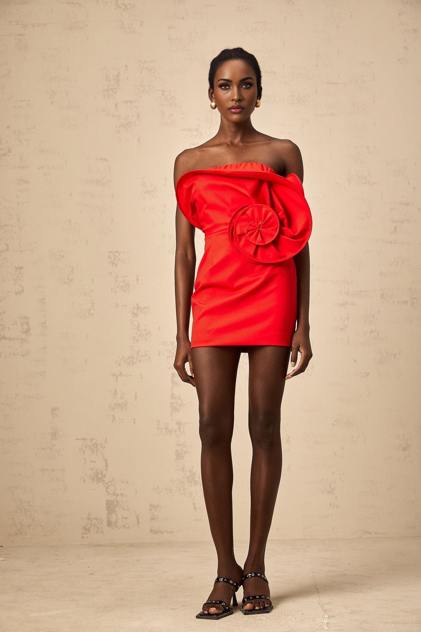 A woman wearing a vibrant red mini dress with an offshoulder neckline featuring a large sculptural rose detail on one shoulder The dress has a fitted silhouette and falls above the knee