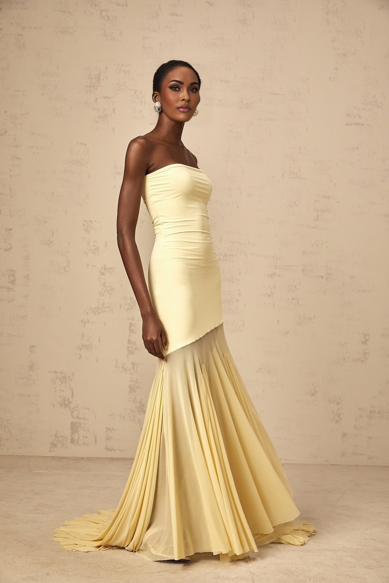 A woman wearing a vibrant yellow offshoulder maxi dress with a sheer tulle overlay standing against a plain background