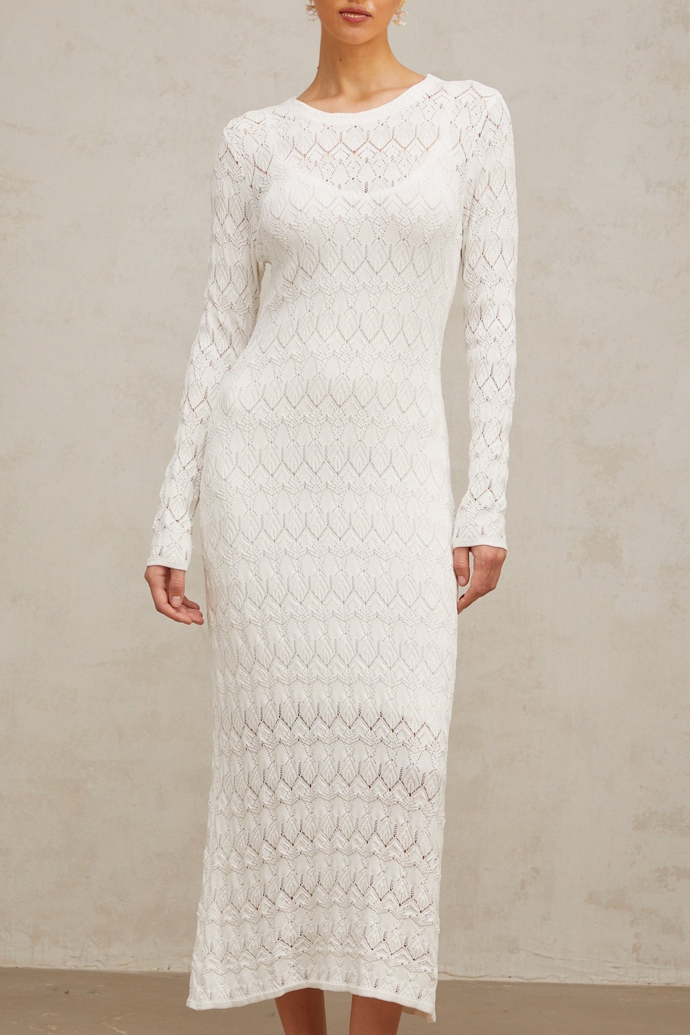 A woman wearing a white crochet knit midi dress with intricate lace patterns standing against a plain background