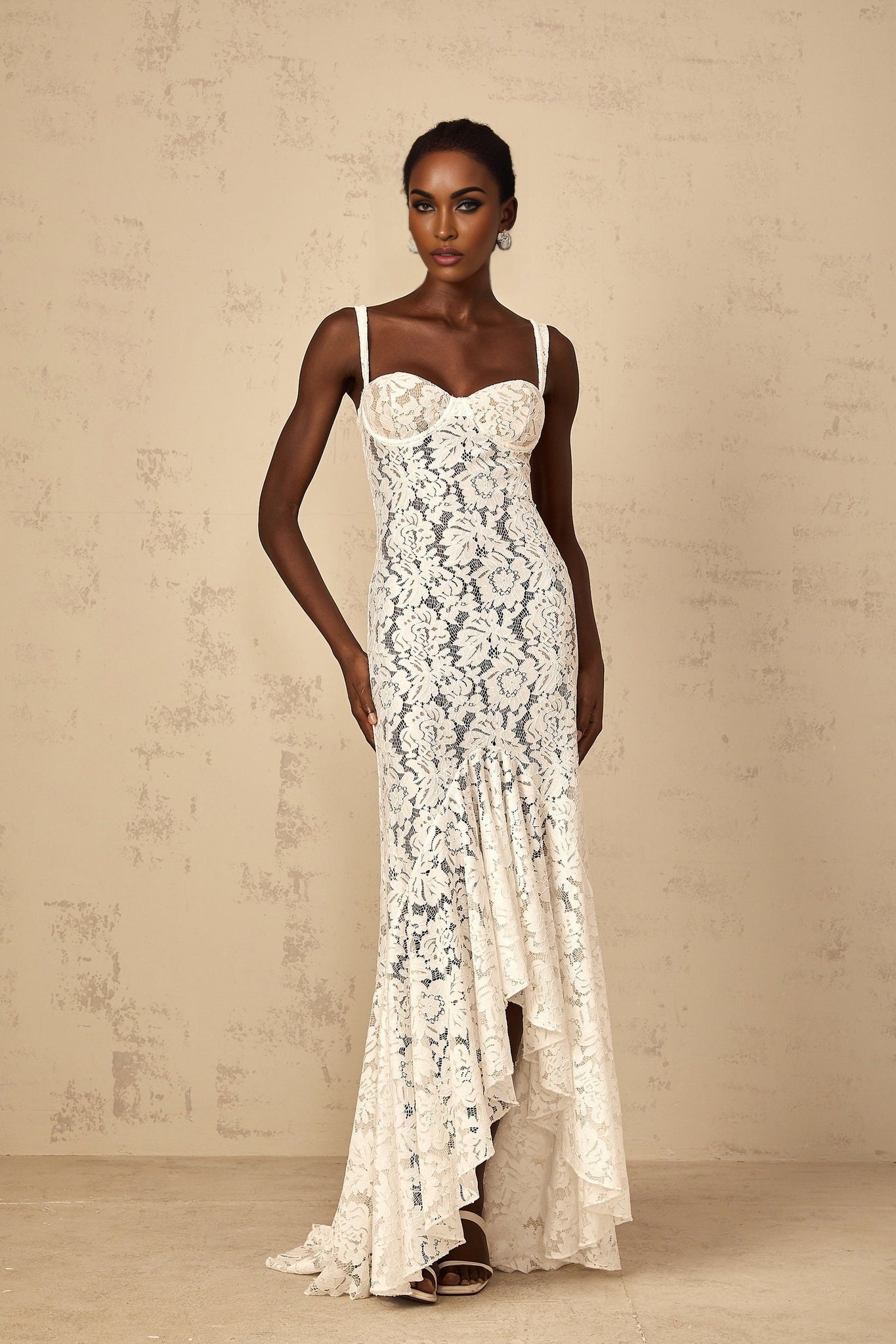 A woman wearing a white floral lace bustier maxi dress with a fitted bodice and flowing skirt standing against a plain background