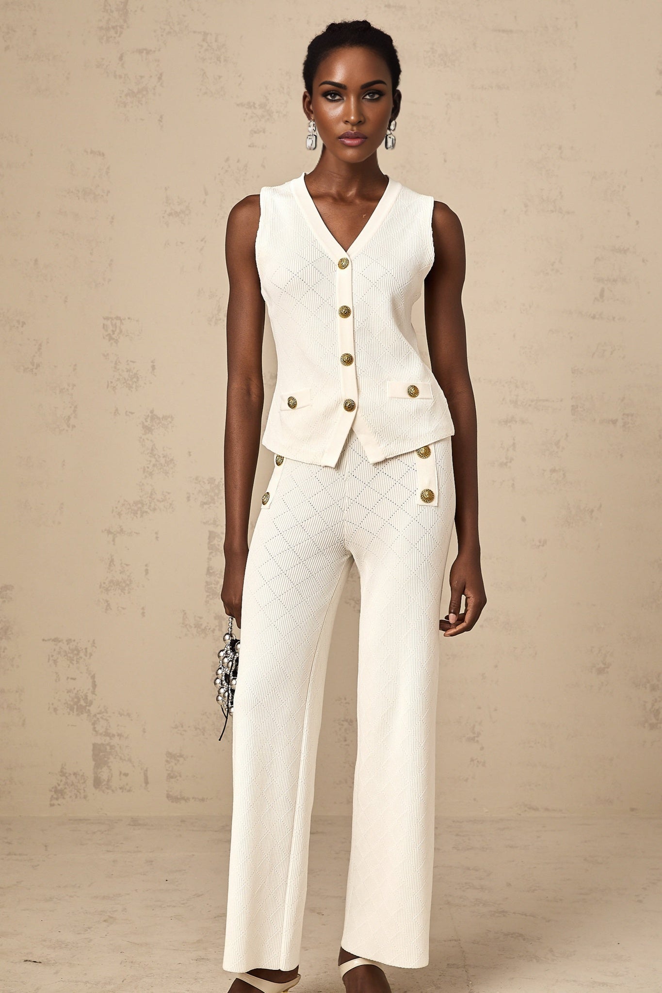 A woman wearing a white knitted matching set consisting of a sleeveless top and loose fitting trousers standing against a plain background