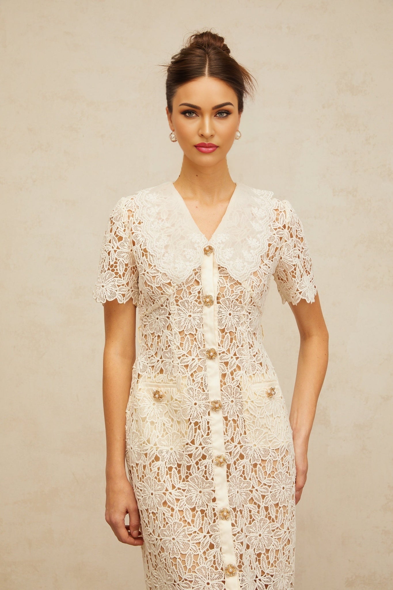 A woman wearing a white lace midi dress with floral patterns and corded details standing against a plain background