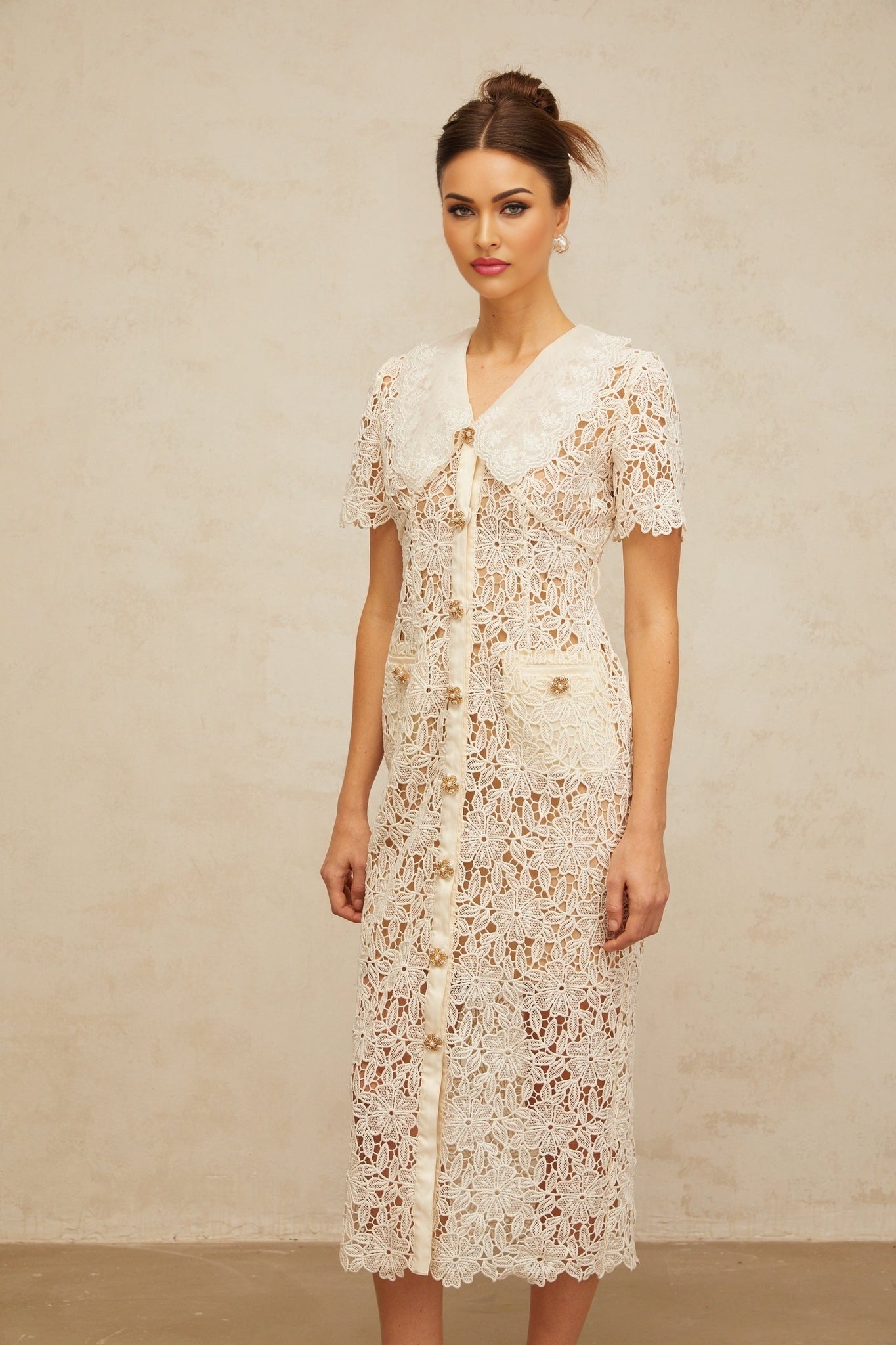 A woman wearing a white lace midi dress with off the shoulder sleeves standing against a plain background