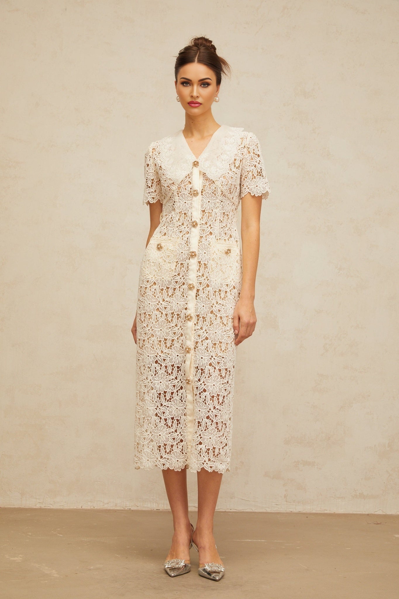 A woman wearing a white lace midi dress with off the shoulder sleeves standing against a plain background