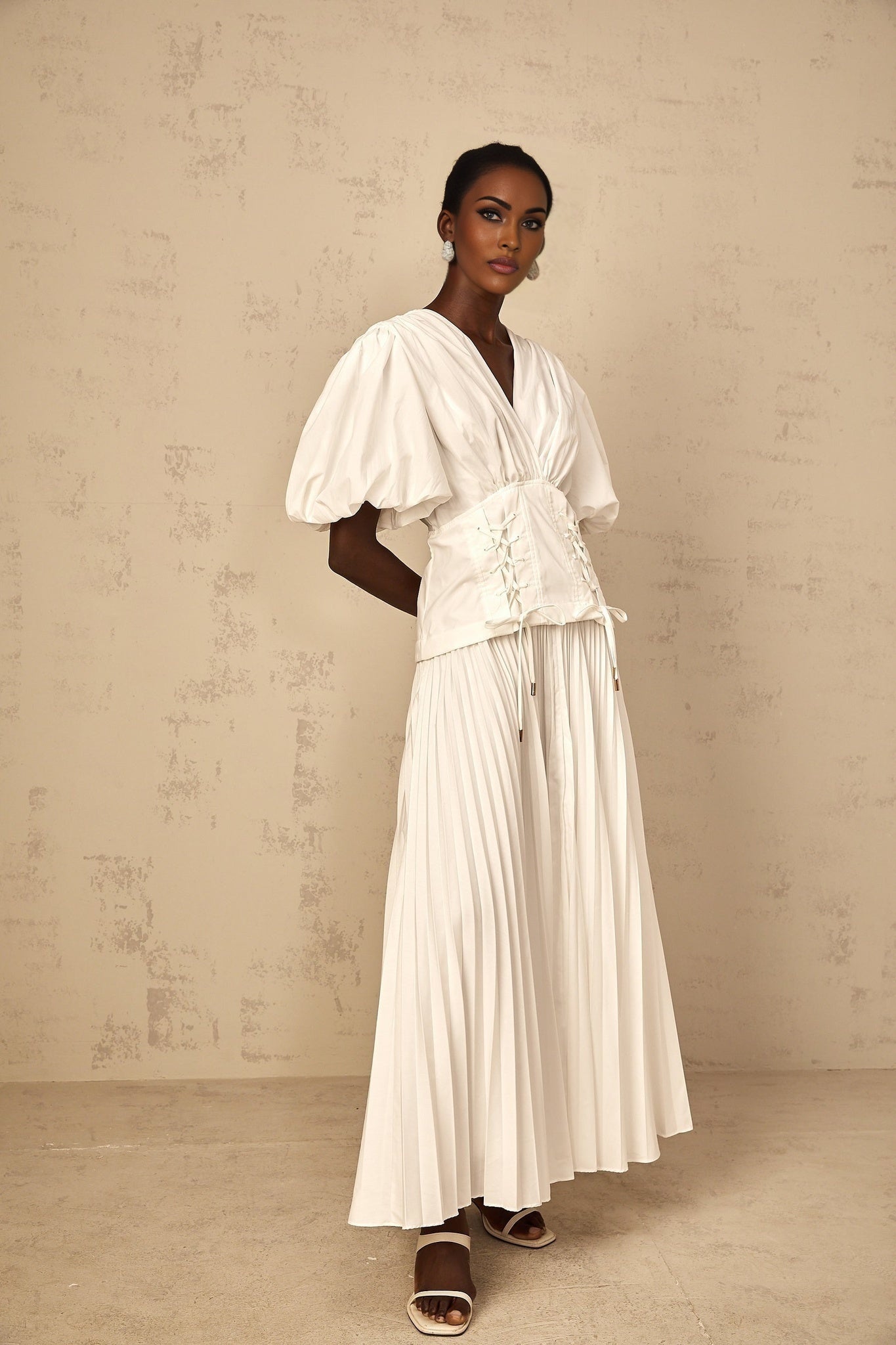 A woman wearing a white lace up pleated maxi dress with long sleeves standing in a relaxed pose