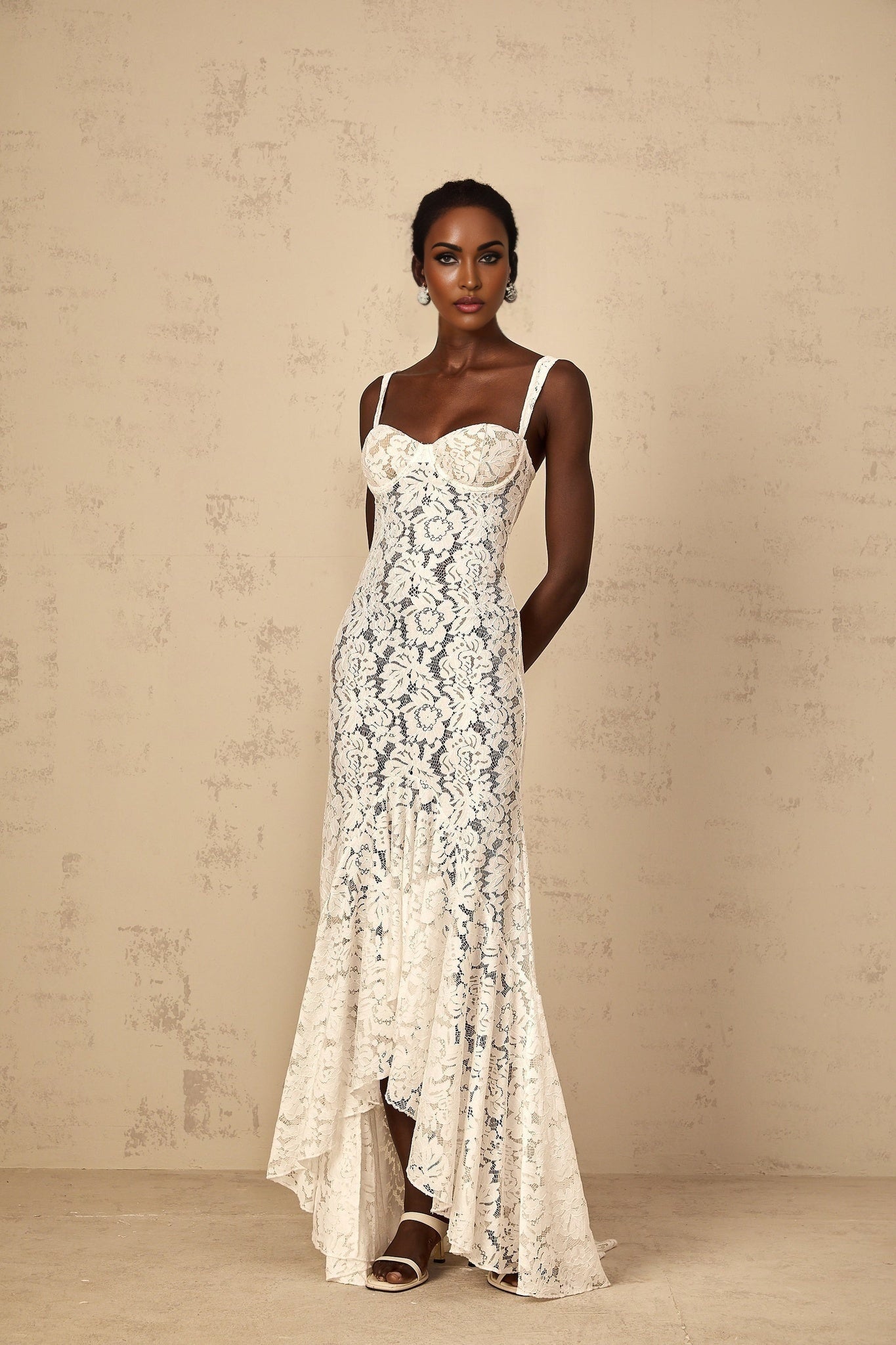 A woman wearing a white maxi dress with floral lace detailing and a bustier bodice standing against a plain background