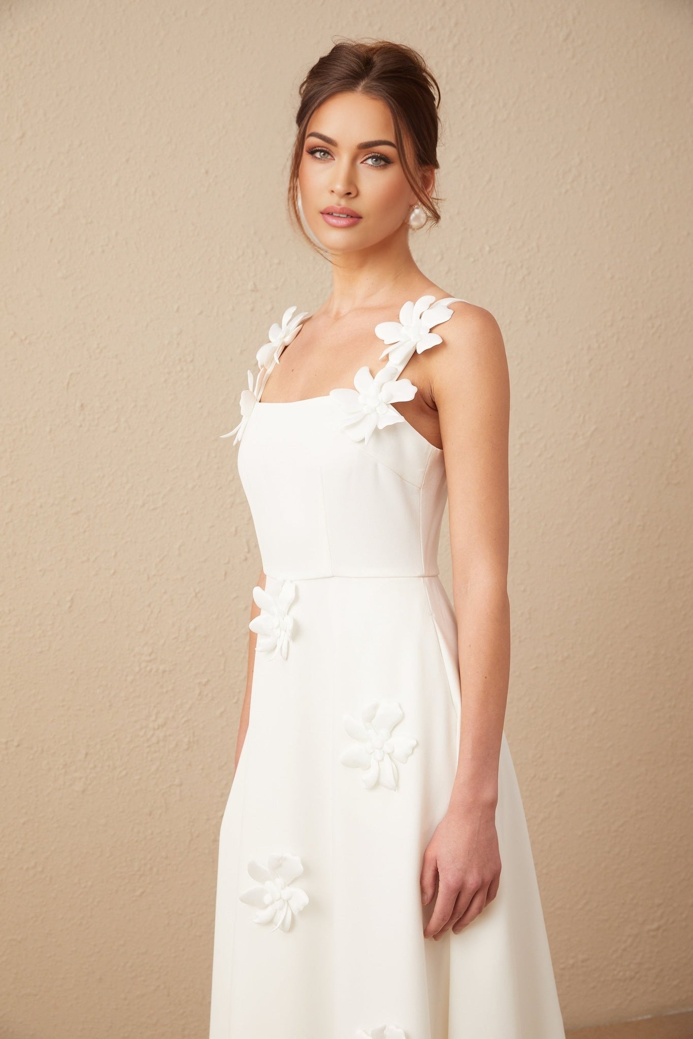 A woman wearing a white midi dress with a square neckline adorned with faux flower details standing against a plain background