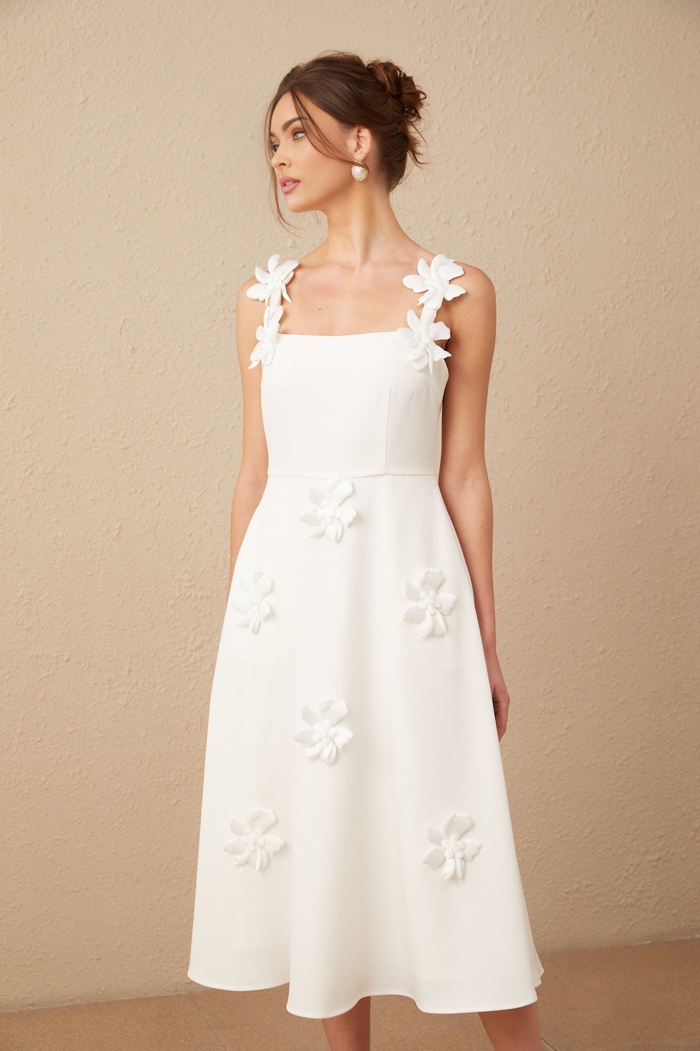 A woman wearing a white midi dress with a square neckline adorned with faux flower details standing against a plain background