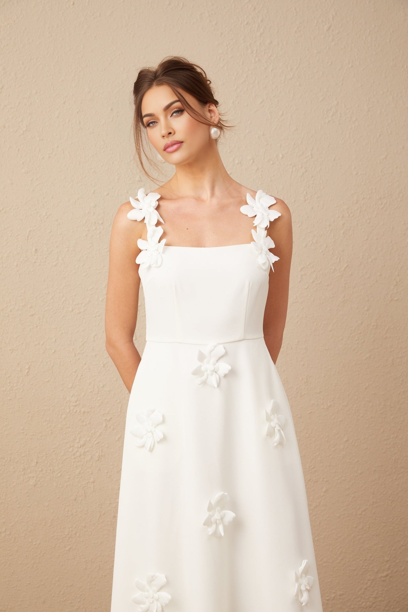 A woman wearing a white midi dress with a square neckline adorned with faux flower embellishments standing against a plain background
