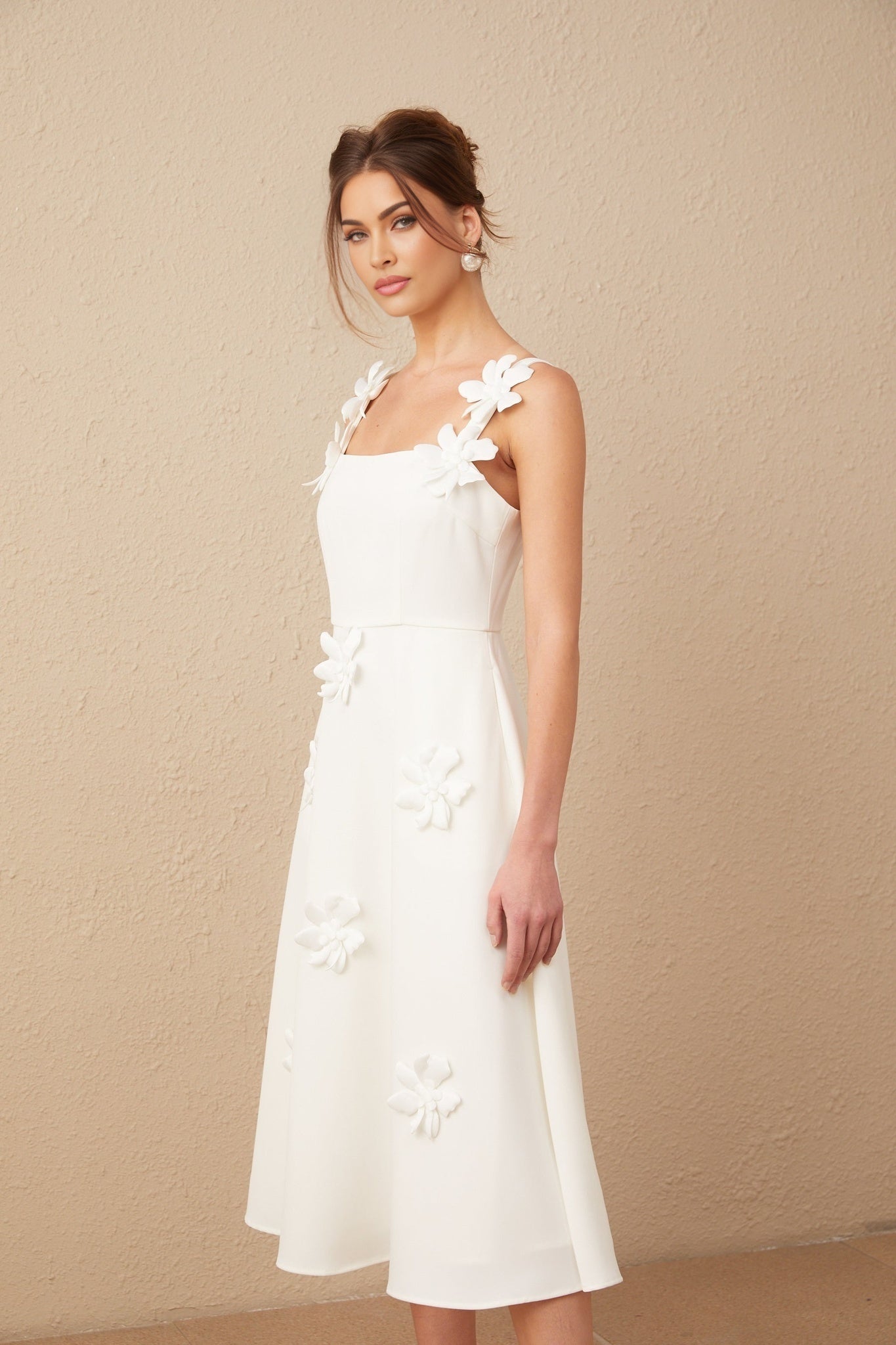 A woman wearing a white midi dress with a square neckline and faux flower embellishments standing in a neutral background