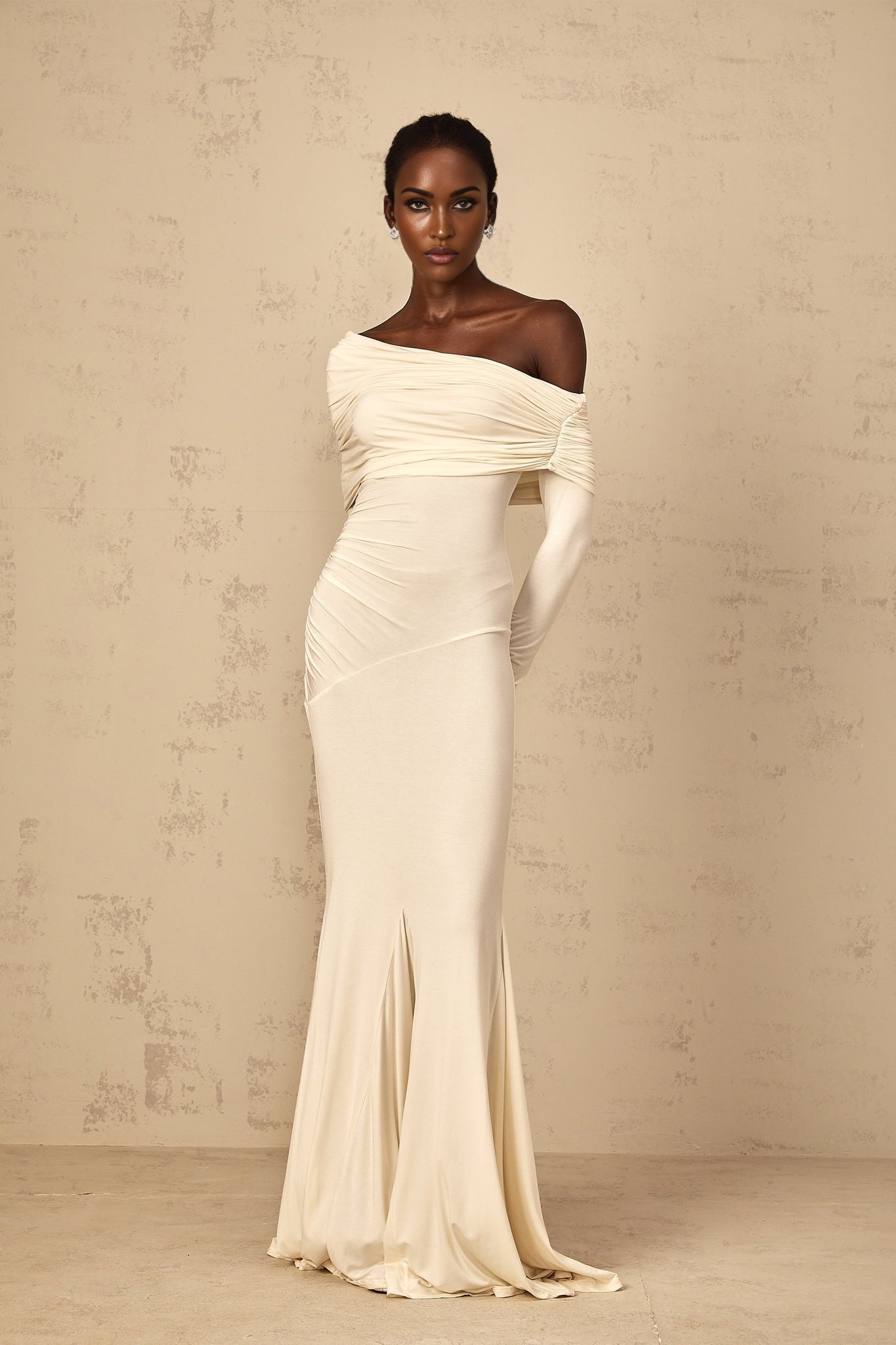 A woman wearing a white offshoulder maxi dress with ruched detailing standing in a relaxed pose against a plain background