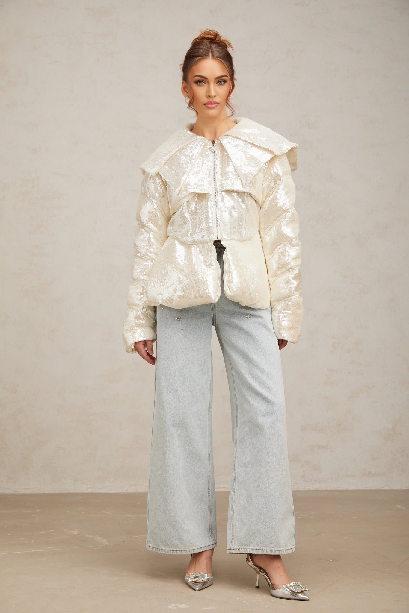 A woman wearing a white puffy down coat covered in sparkling sequins standing against a plain background