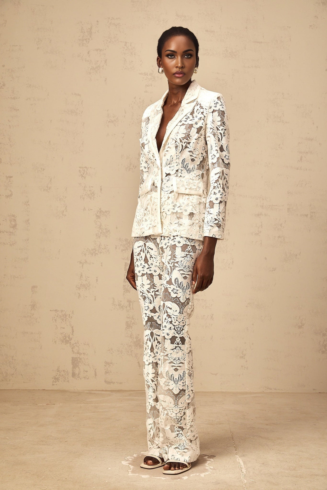 A woman wearing a white sequined blazer and matching trousers featuring floral embroidery on tulle fabric The set has a glamorous and elegant appearance with a shimmering effect from the sequins