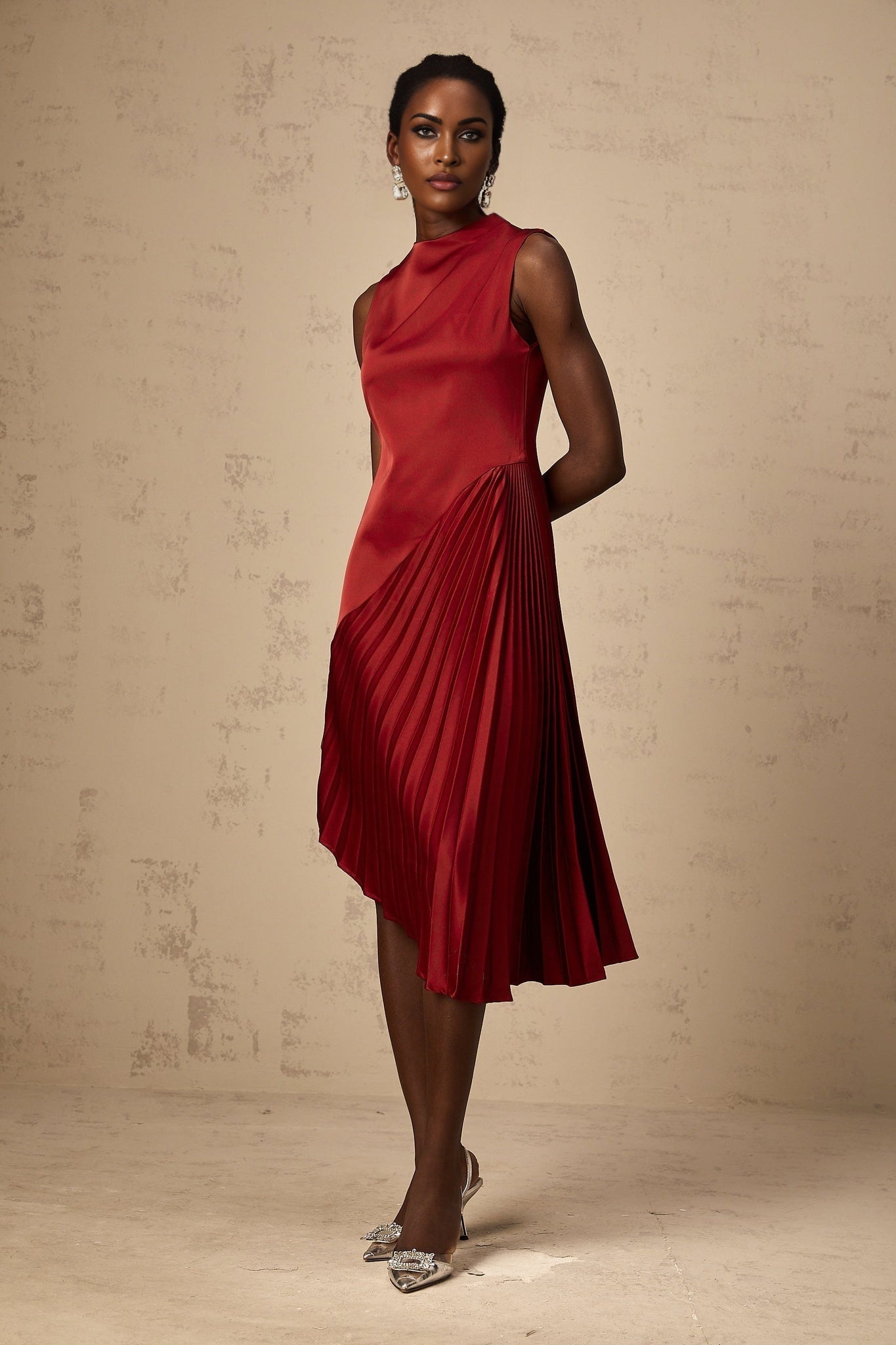 A woman wearing a wine red midi dress with an asymmetric neckline and pleated skirt standing against a neutral background
