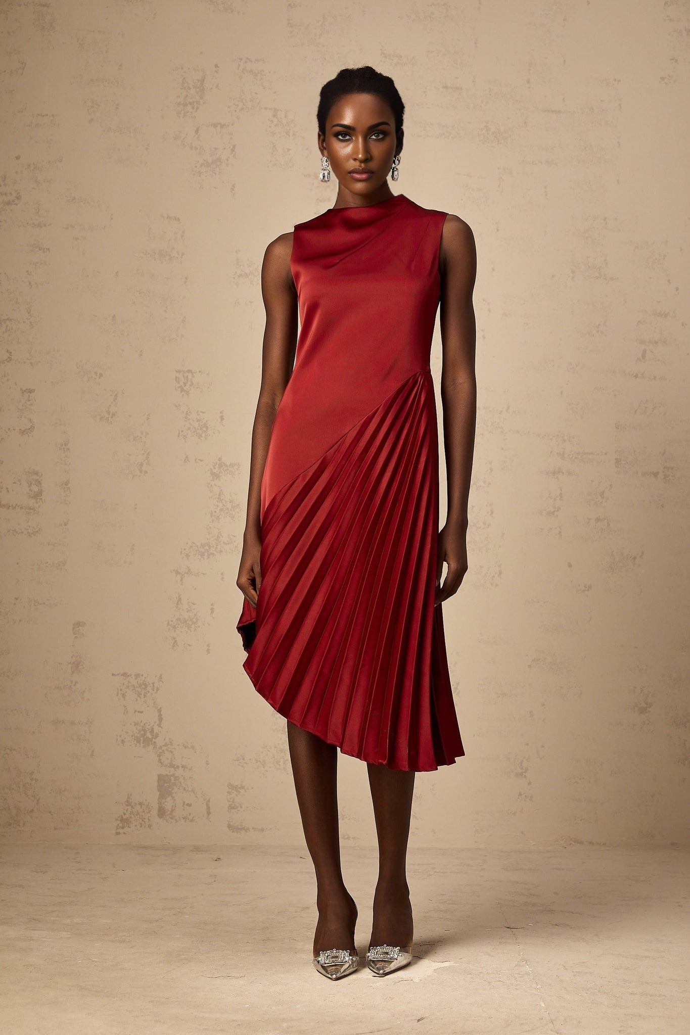 A woman wearing a wine red midi dress with an asymmetric neckline and pleated skirt standing against a plain background