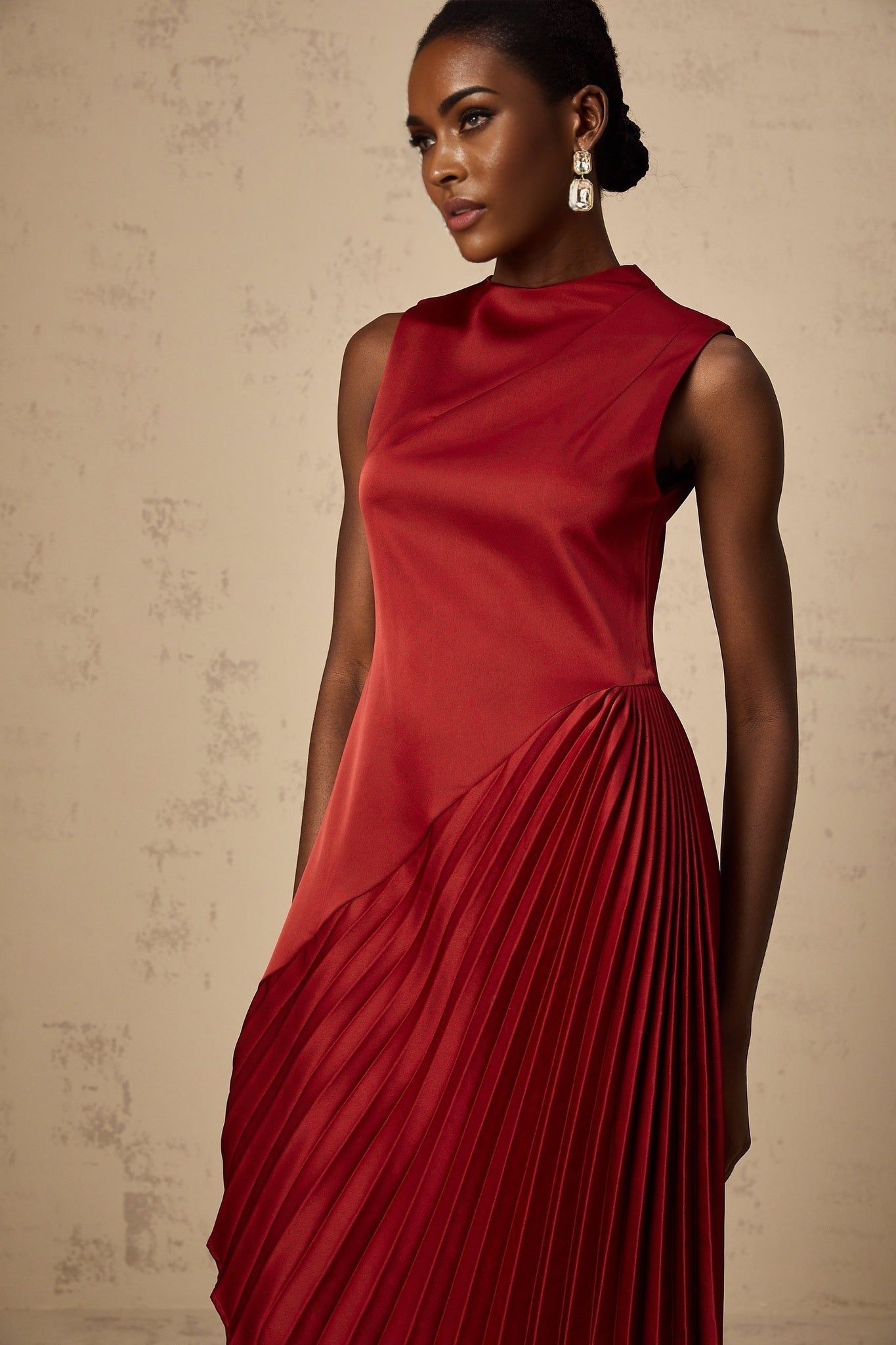 A woman wearing a wine red midi dress with an asymmetric neckline and pleated skirt standing against a plain background