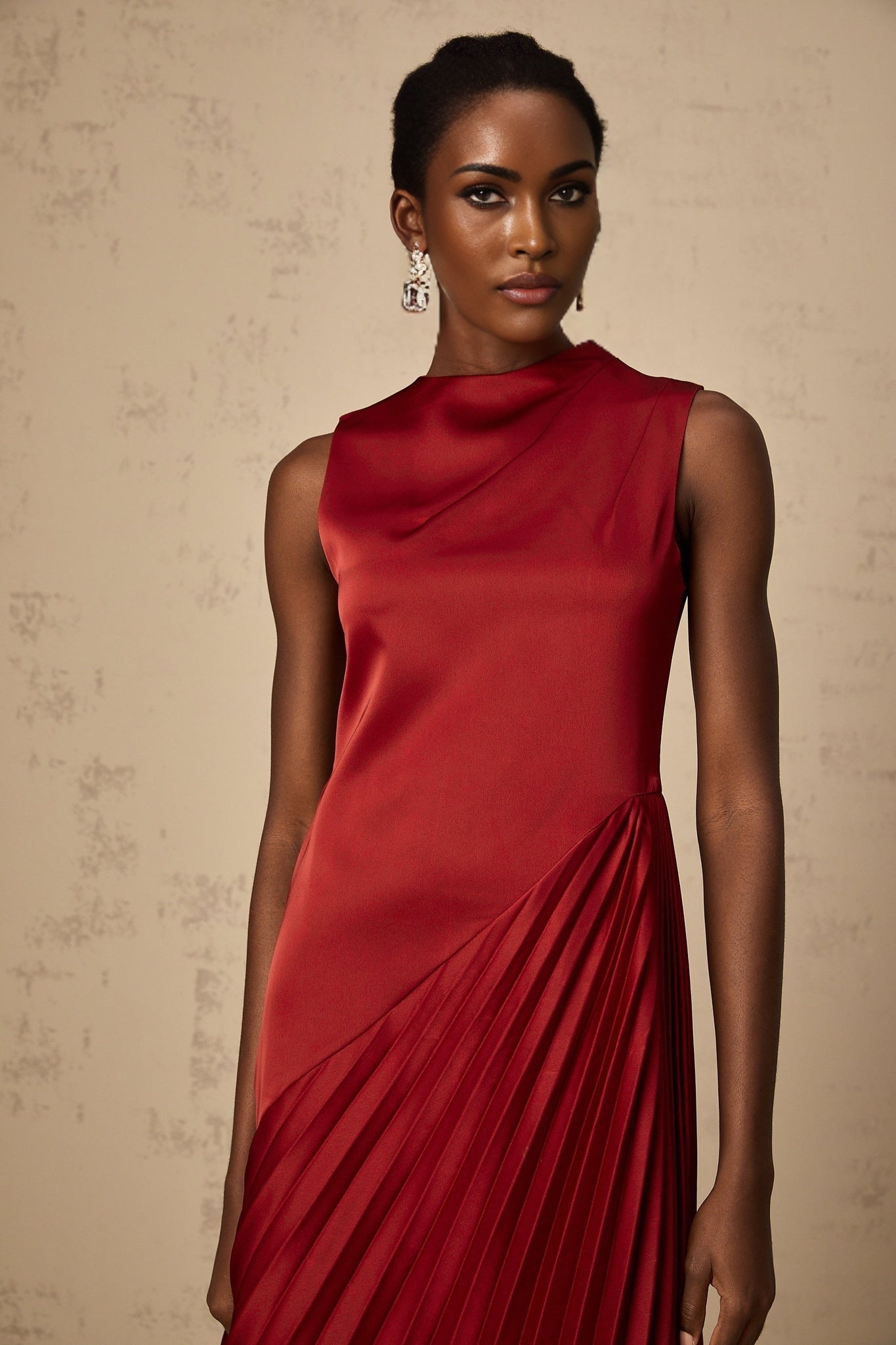 A woman wearing a wine red midi dress with an asymmetric neckline and pleated skirt standing against a plain background