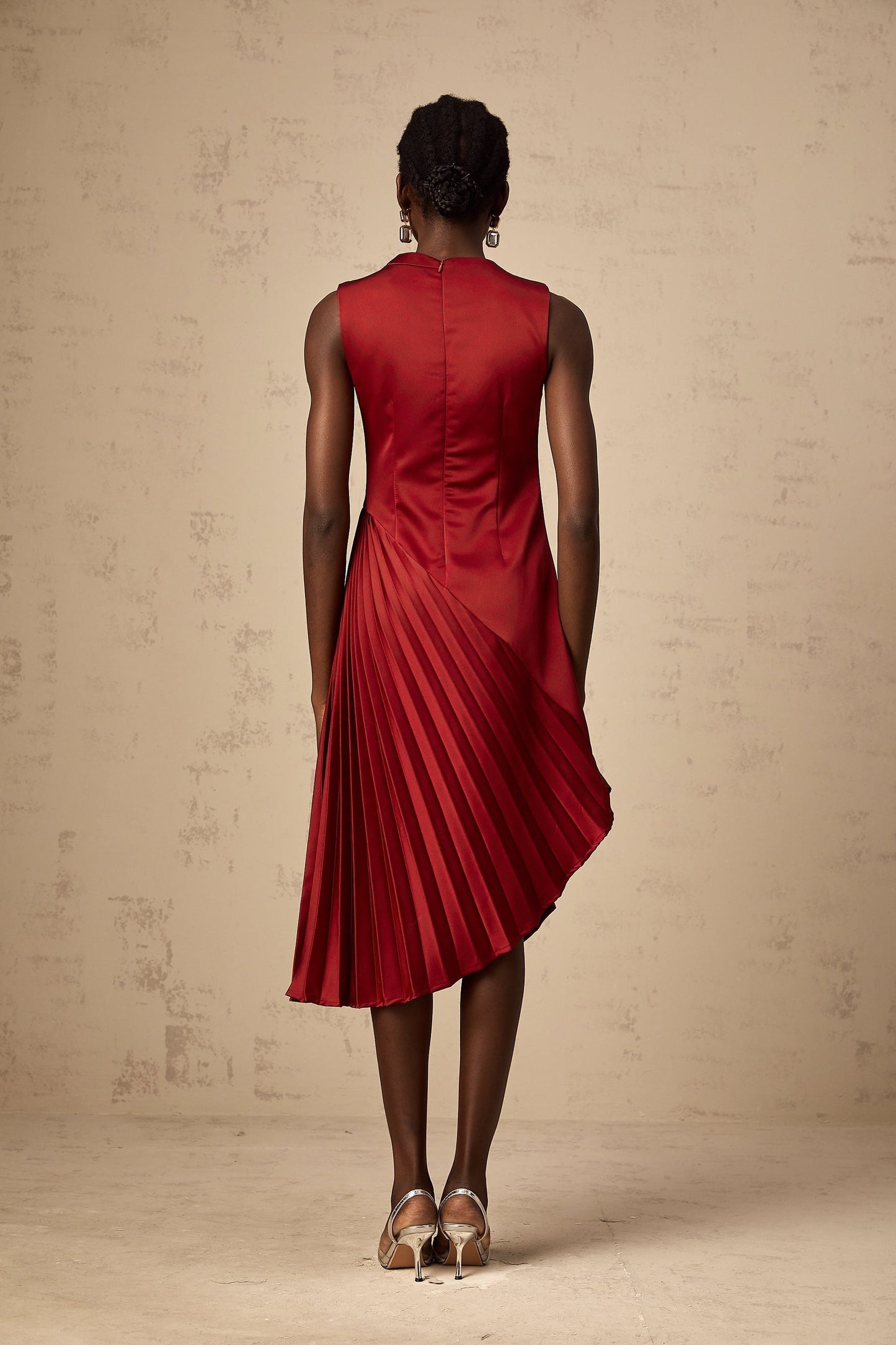A woman wearing a wine red midi dress with an asymmetric neckline and pleated skirt standing against a plain background