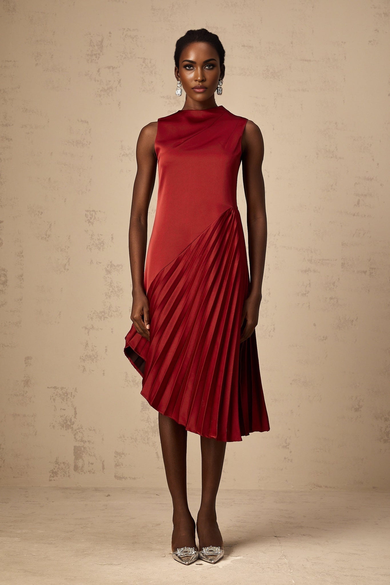 A woman wearing a wine red midi dress with an asymmetric neckline and pleated skirt standing against a plain background