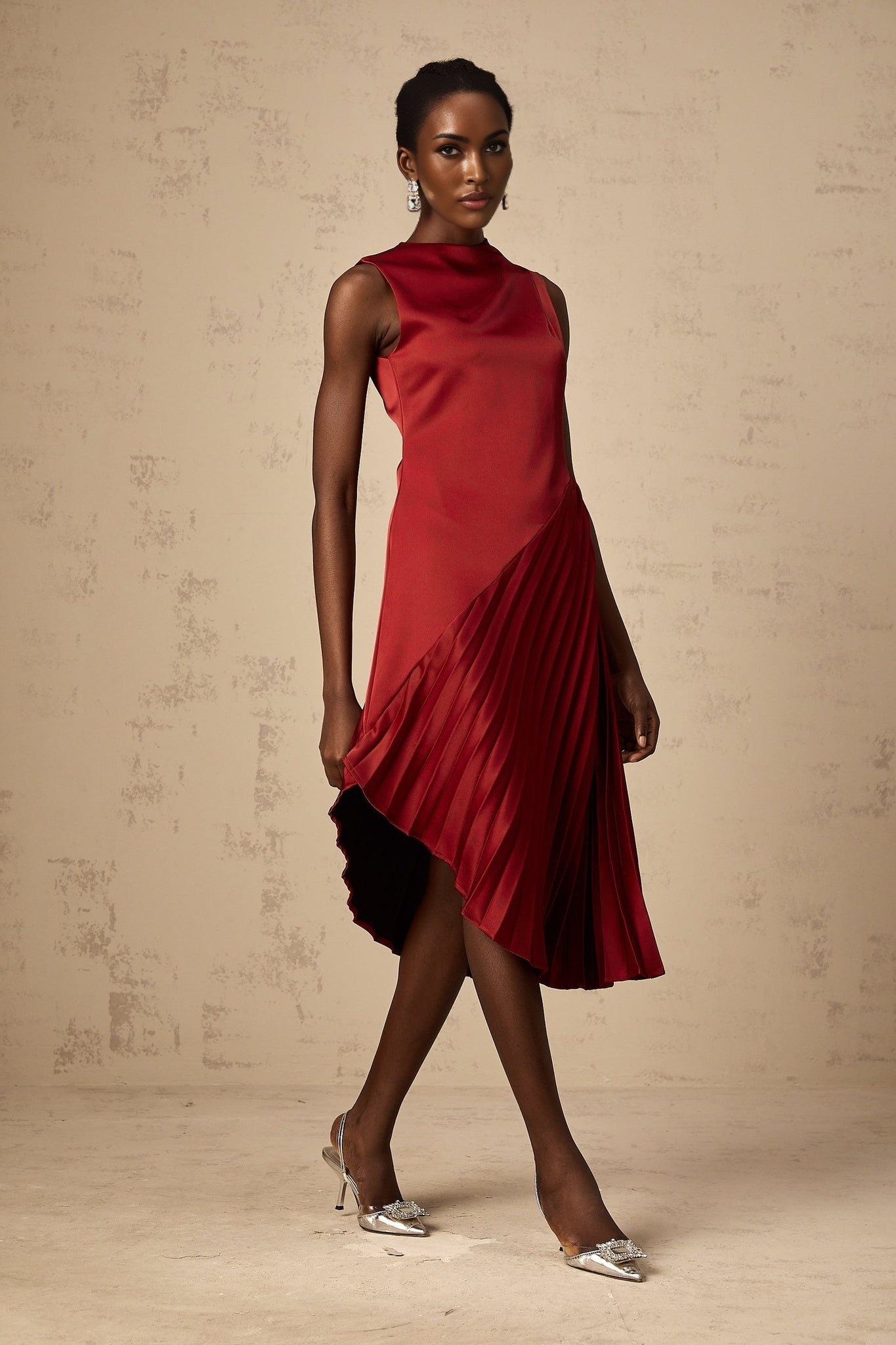 A woman wearing a wine red midi dress with an asymmetric neckline and pleated skirt standing against a plain background