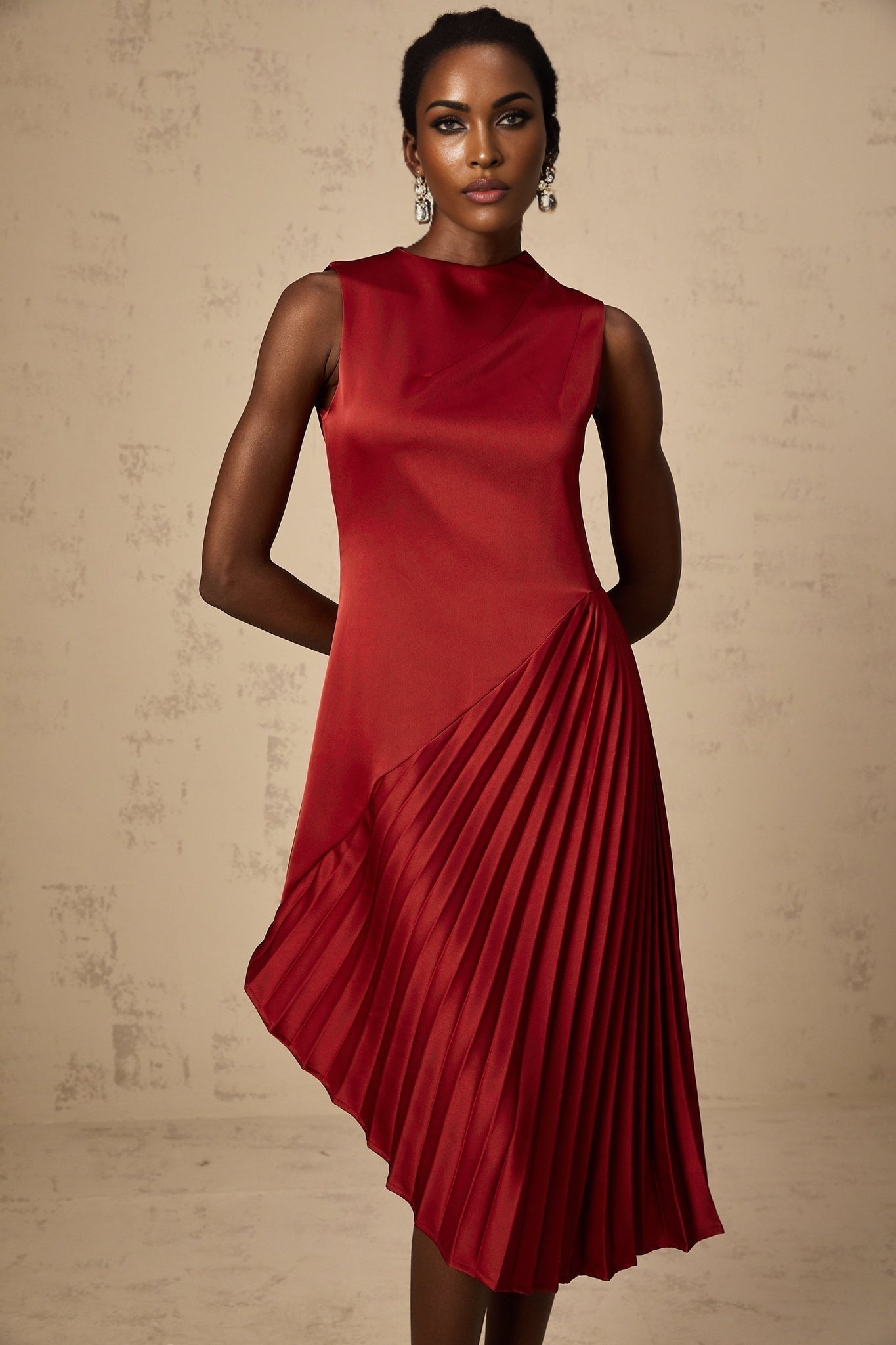 A woman wearing a wine red midi dress with an asymmetric neckline and pleated skirt standing in a neutral setting