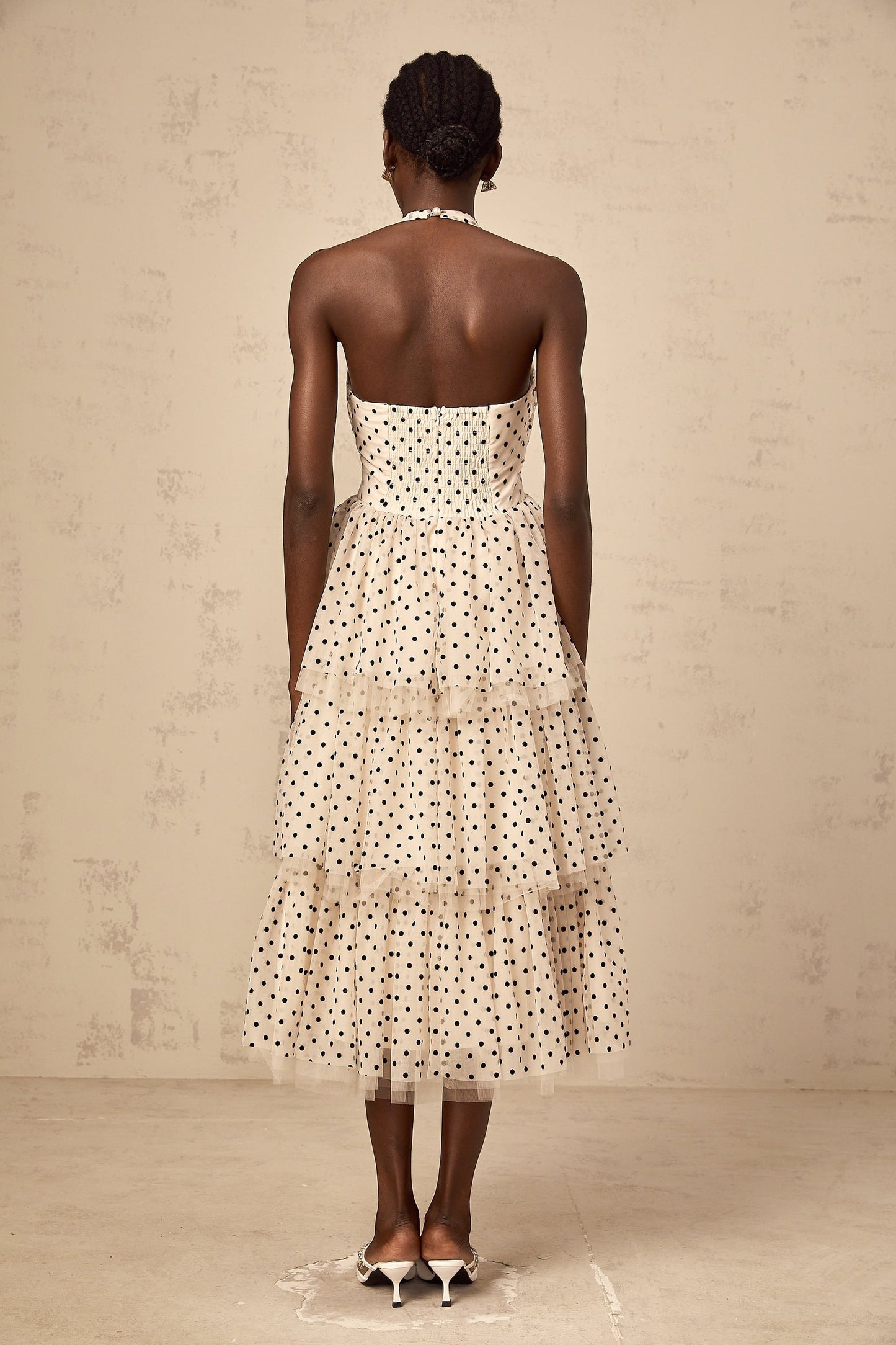 A woman wearing an ivory colored midi dress with polka dots and tiered layers standing in a neutral background