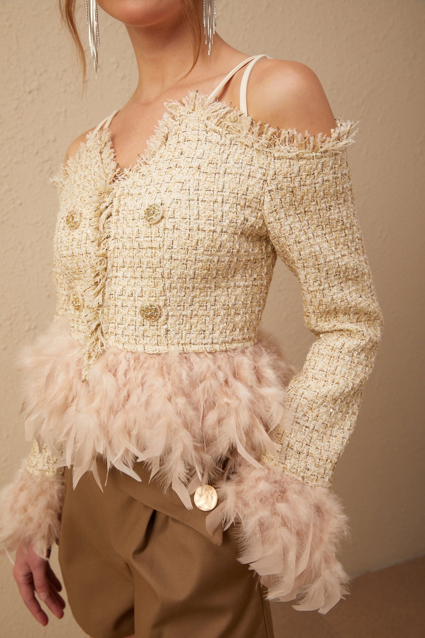 A woman wearing an offshoulder tweed top with feather trim around the neckline The top is in a light color and has a textured tweed fabric The model has her hands on her hips showing off the flattering cut of the garment