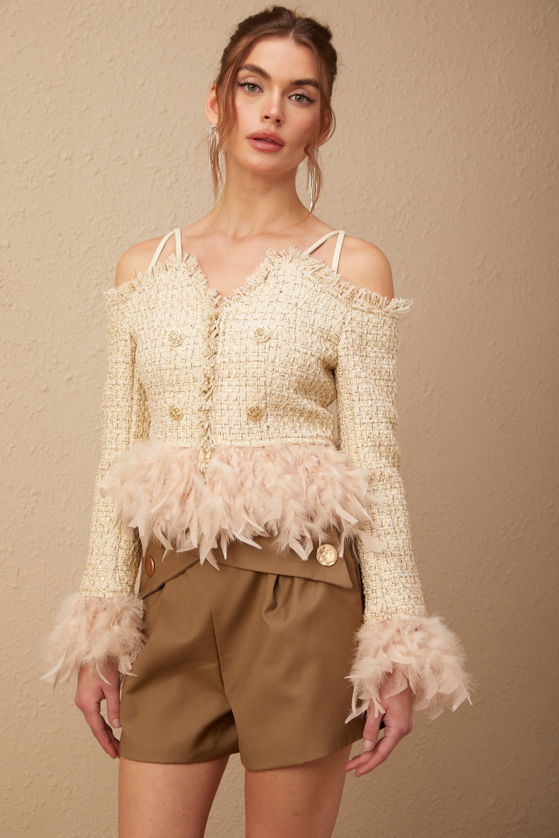 A woman wearing an offshoulder tweed top with feather trim around the neckline The top is light colored and has a textured tweed pattern The model has her arms at her sides showing the shirts offshoulder design