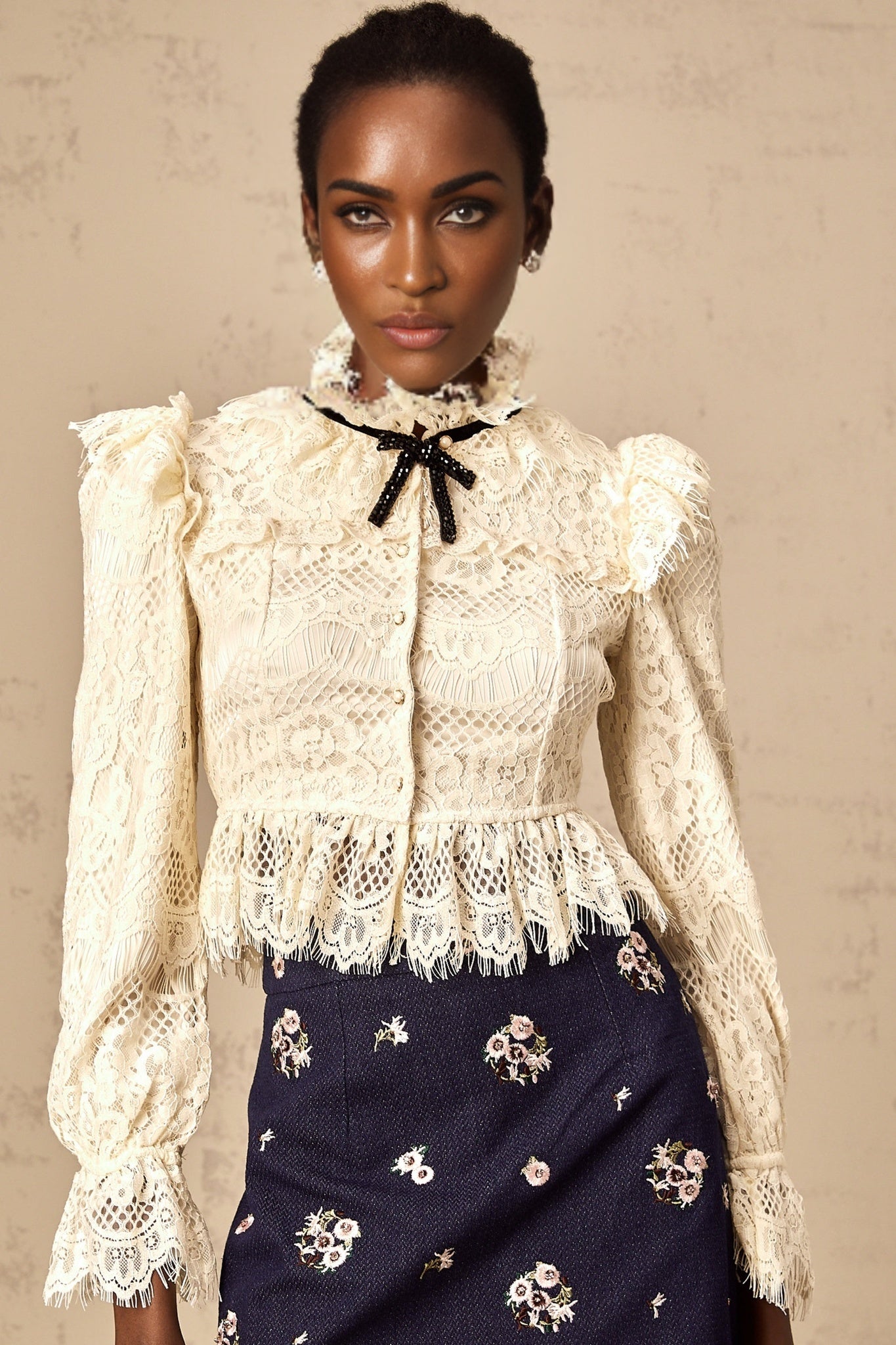 A woman wearing an offwhite blouse with floral lace detailing and a bowknot accent at the neckline