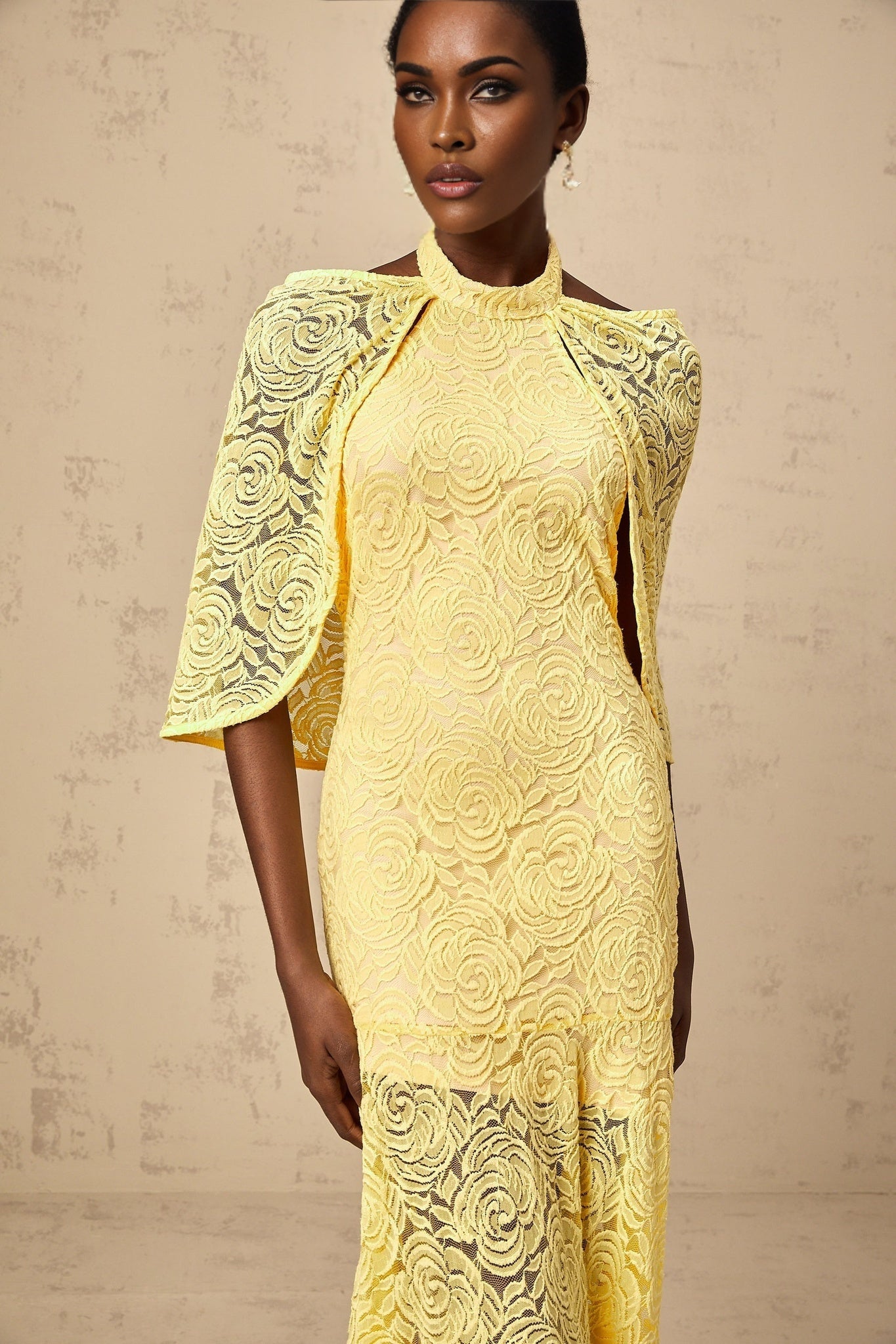 A yellow maxi dress with a cape design made of chantilly lace featuring a long flowing silhouette and delicate floral patterns